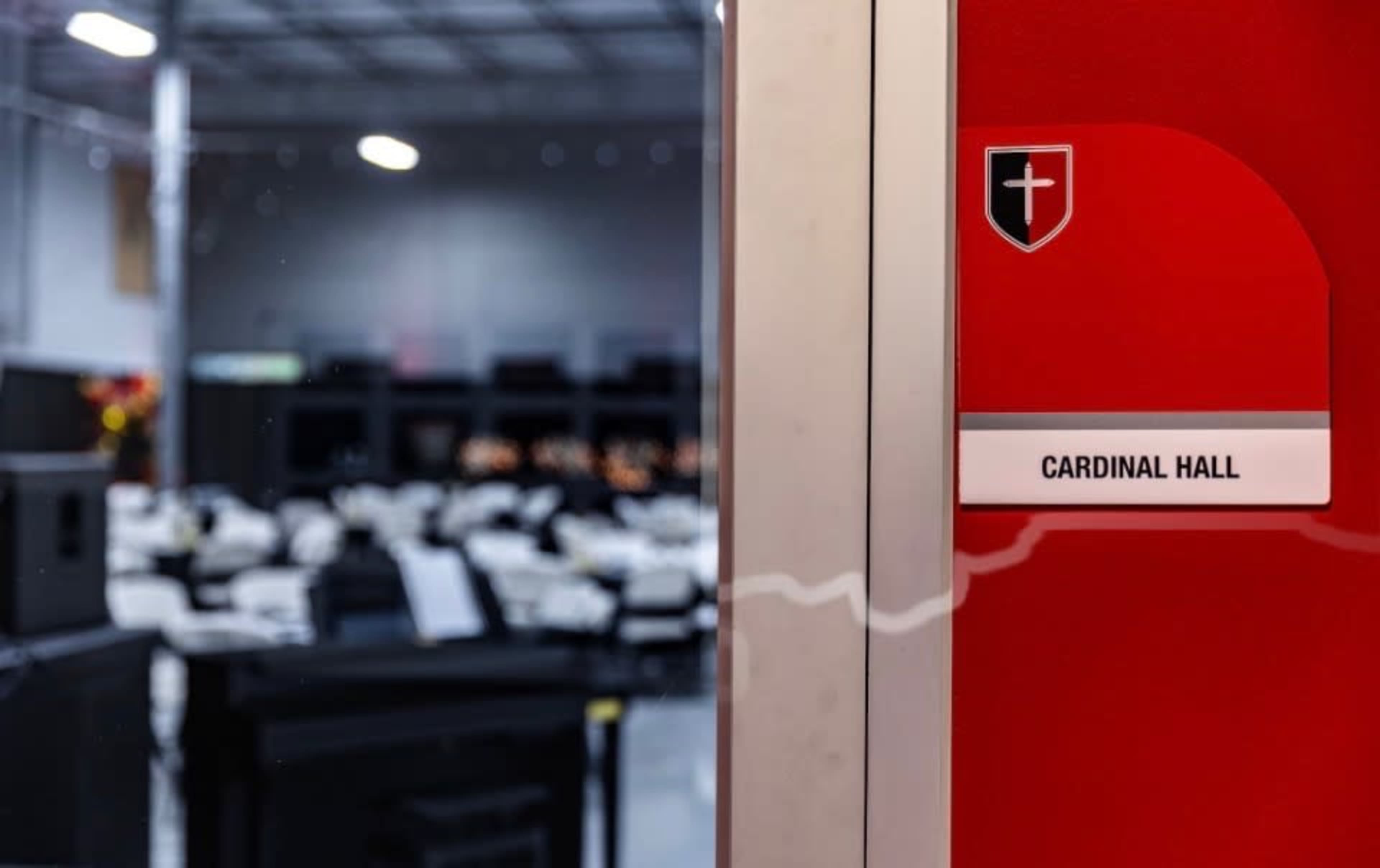 A red door labeled "Cardinal Hall" stands open, revealing a spacious interior with rows of seating in a well-lit environment.