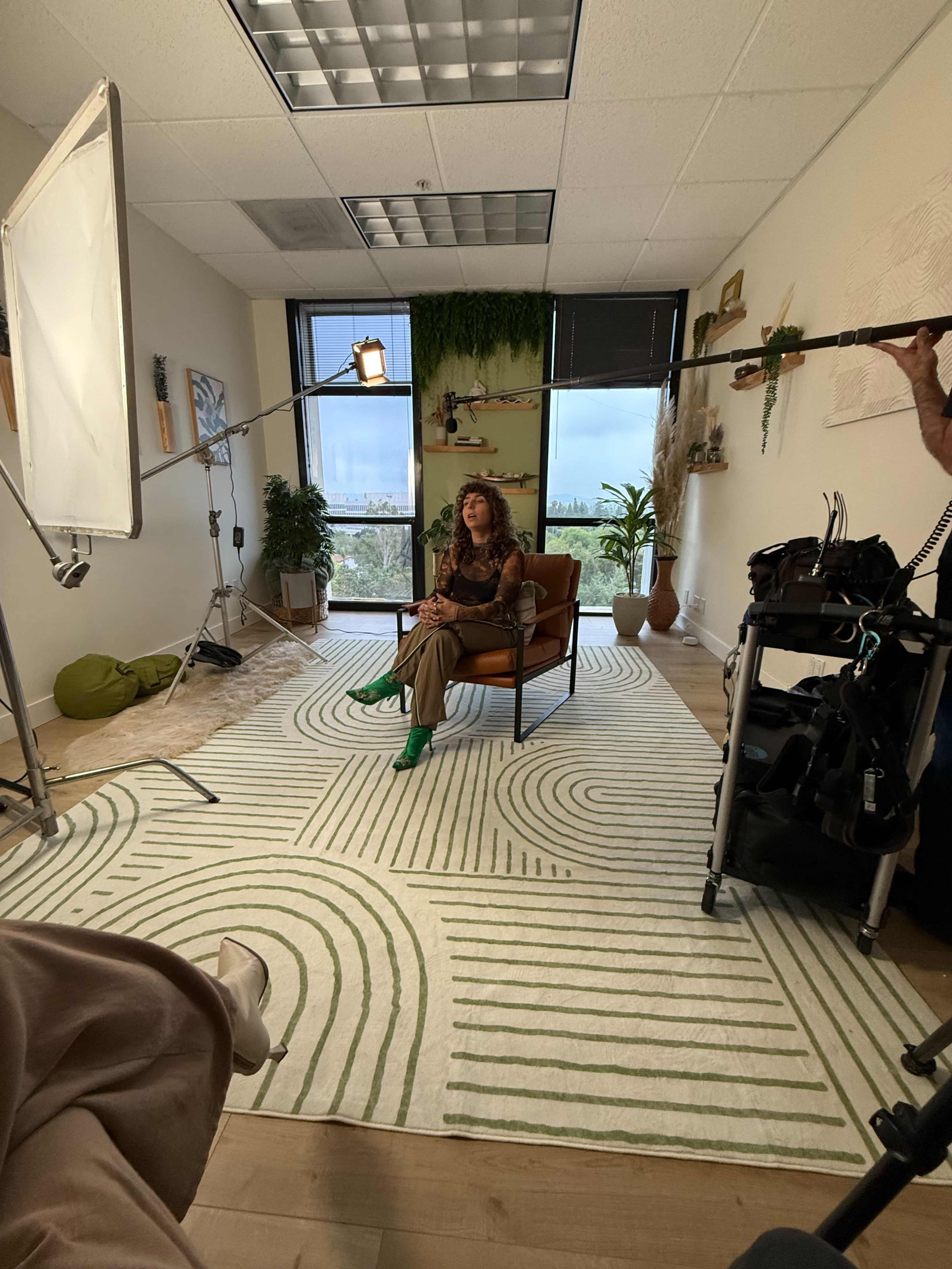 A woman sits in a brown chair in a well-lit room decorated with plants and a green and white rug while being filmed.