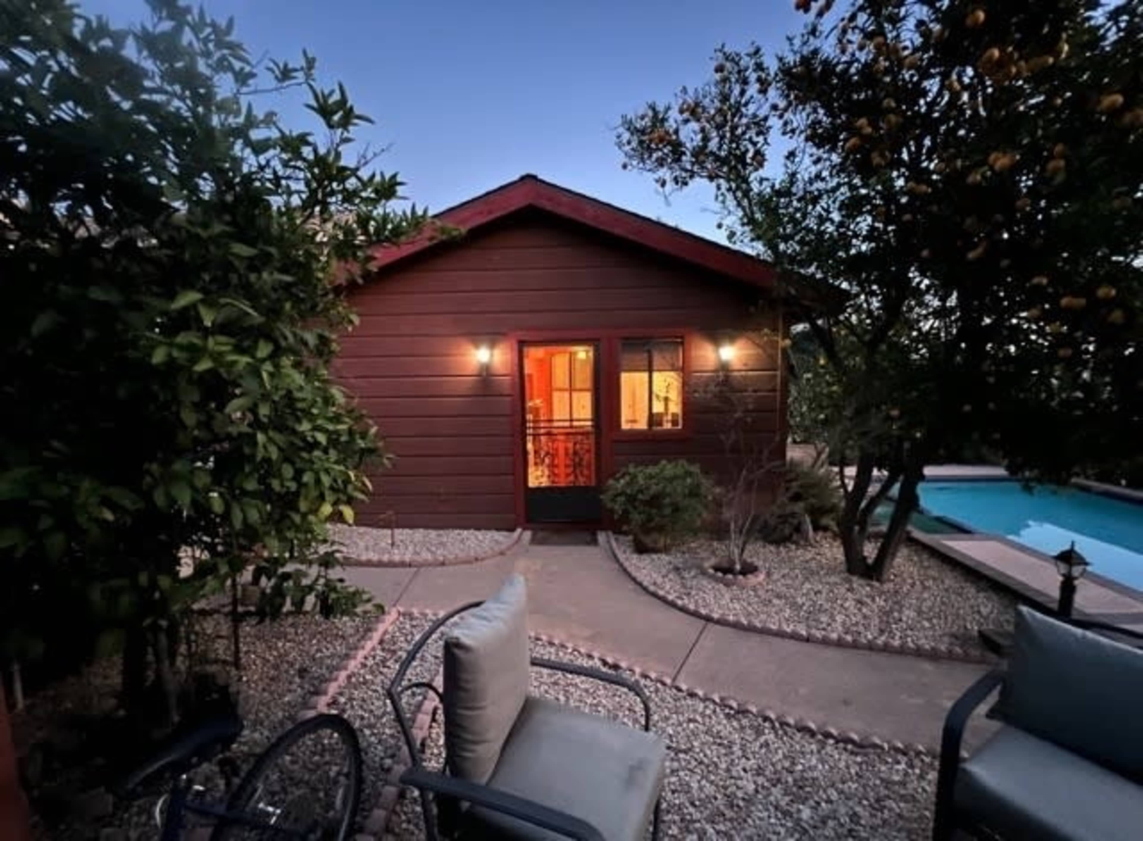 A wooden cabin with lit windows is situated beside a pool, surrounded by trees and a gravel pathway.
