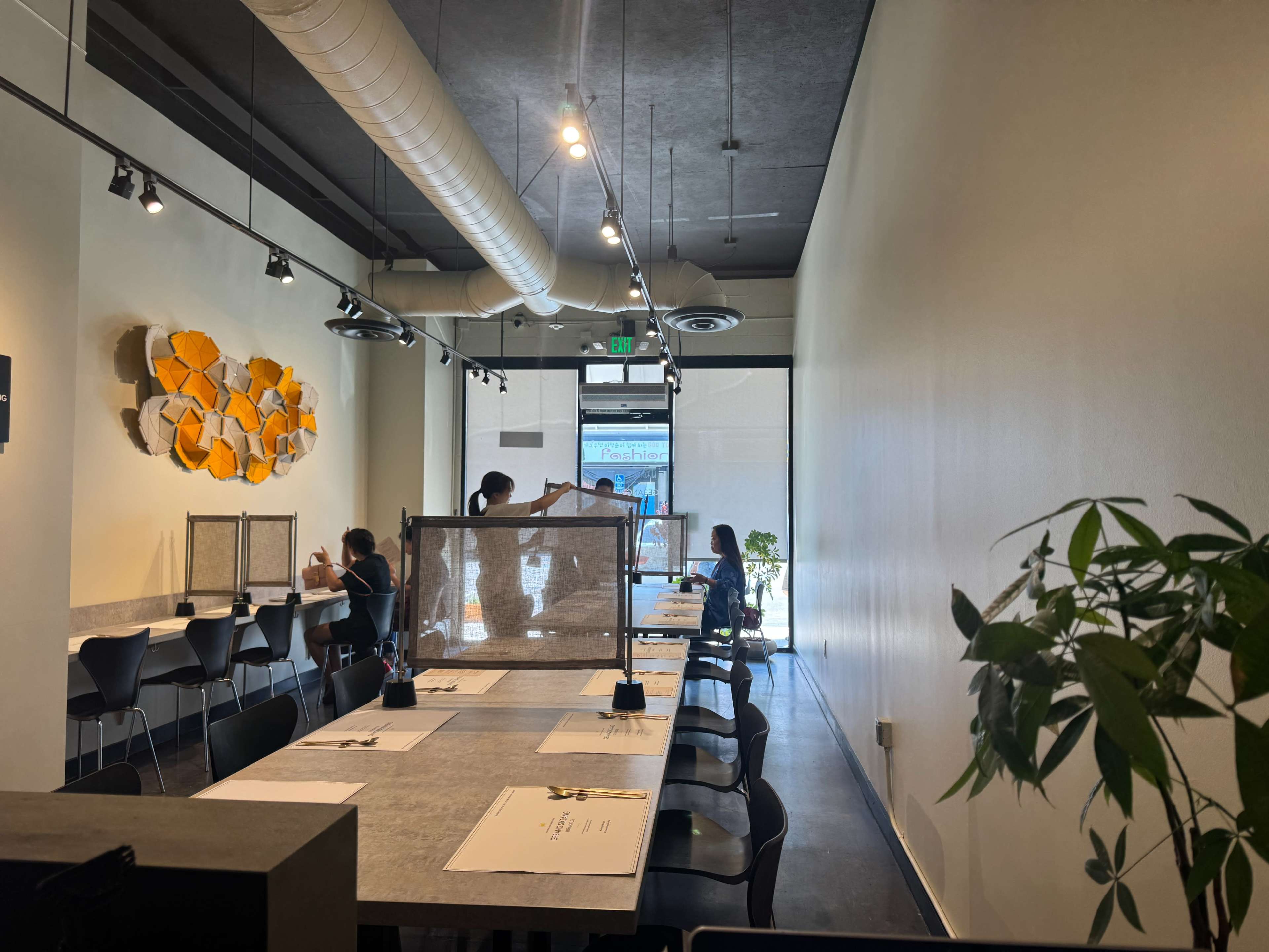 The image shows a modern restaurant interior with tables arranged along the walls, featuring dividers for social distancing, and patrons seated at tables near the entrance.