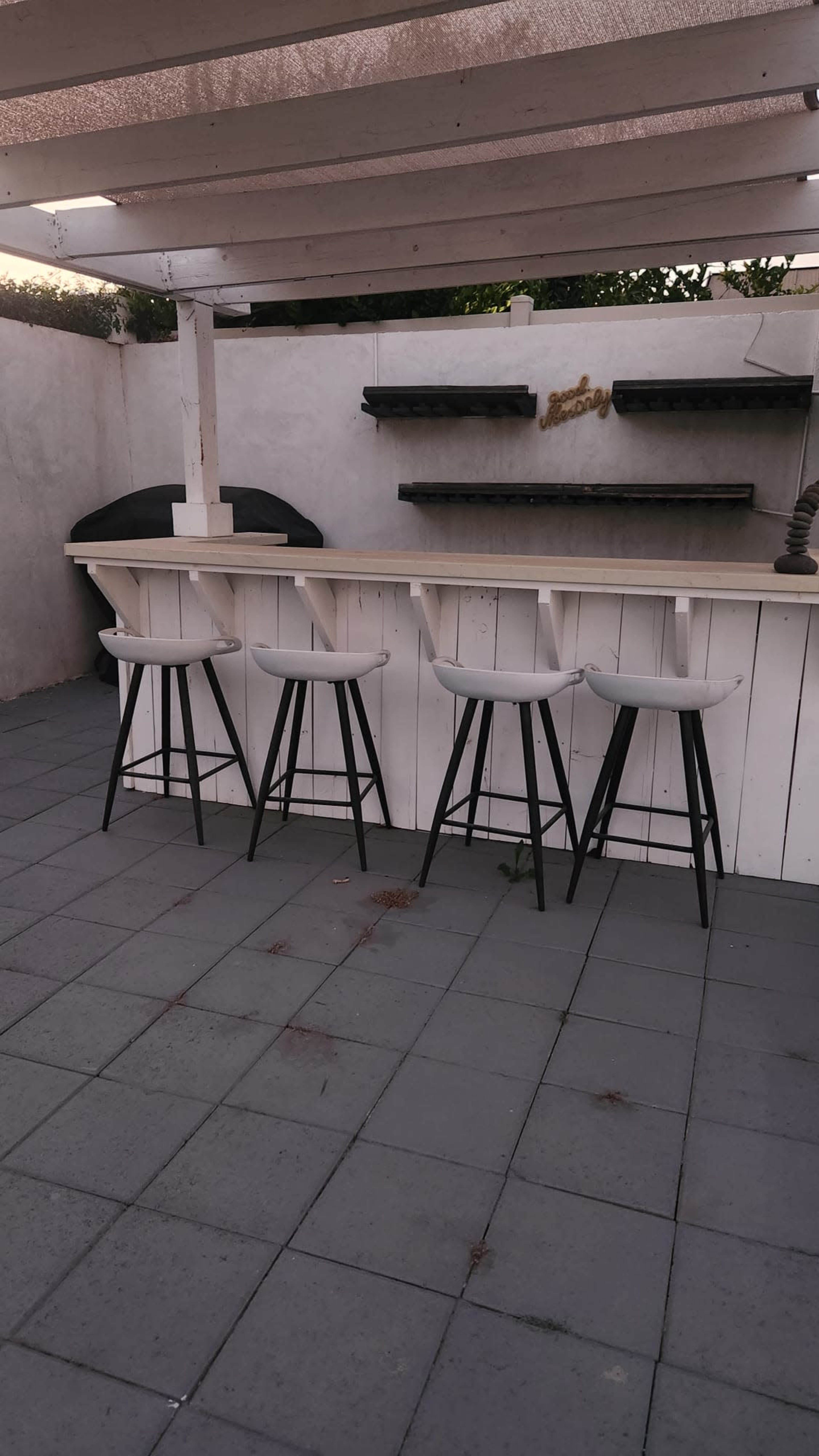 The image shows a patio area with a white bar counter featuring four white stools and a bare wall in the background with shelves.
