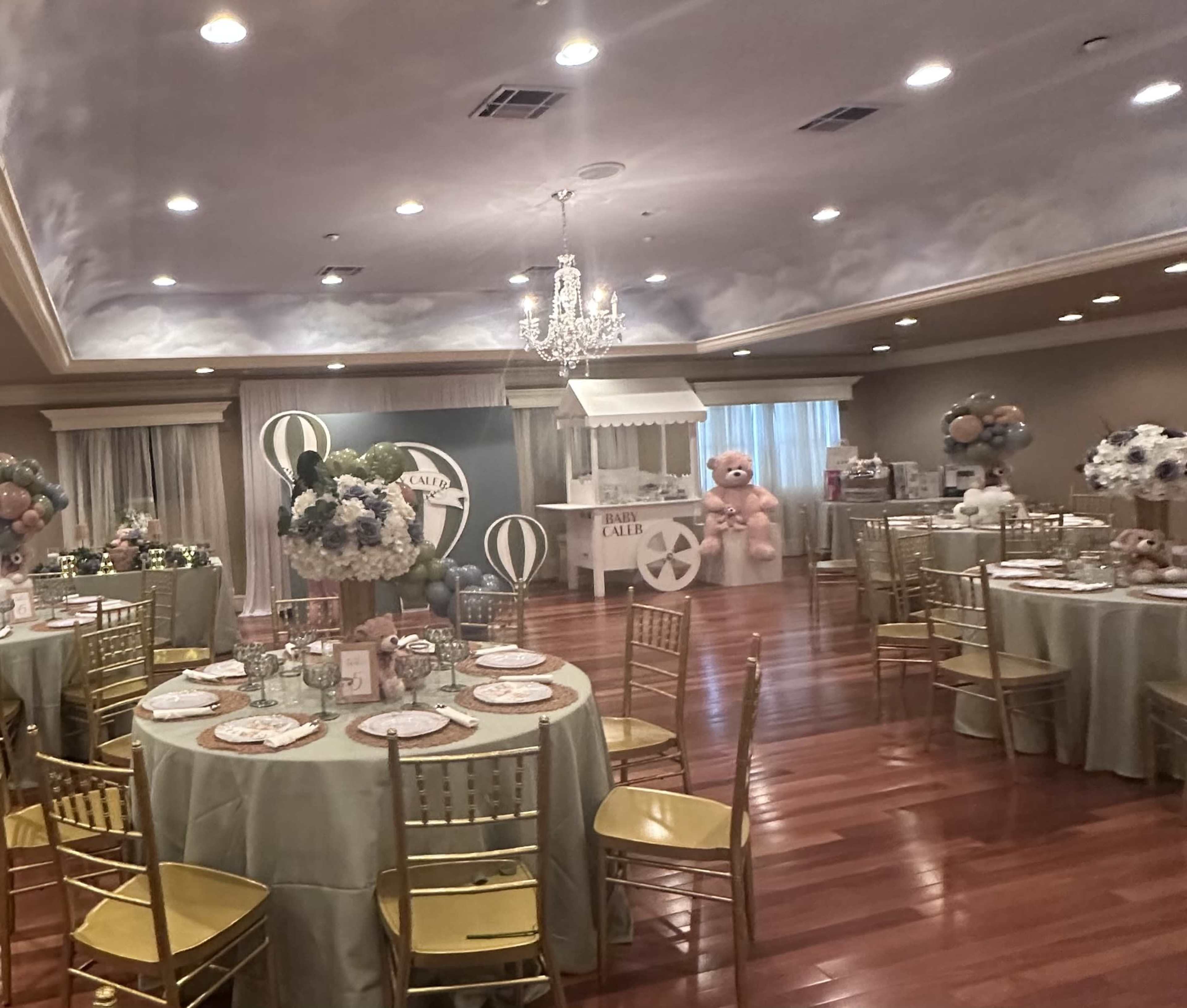 The image shows a decorated event space featuring round tables with green tablecloths, gold chairs, and a dessert station adorned with balloons and floral arrangements.