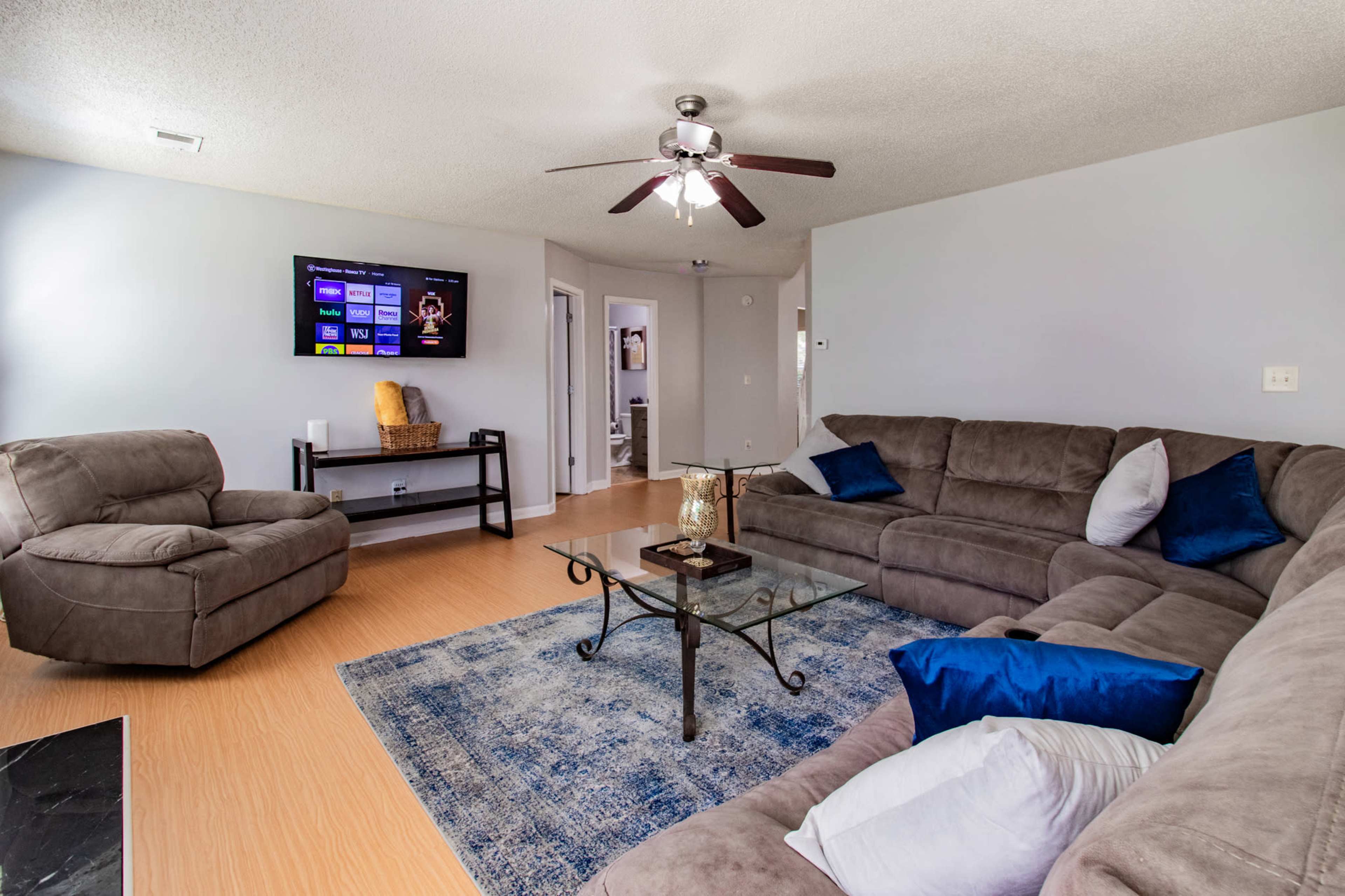 The living room features a sectional sofa, a glass coffee table, and a television mounted on the wall, with a blue area rug on the floor.