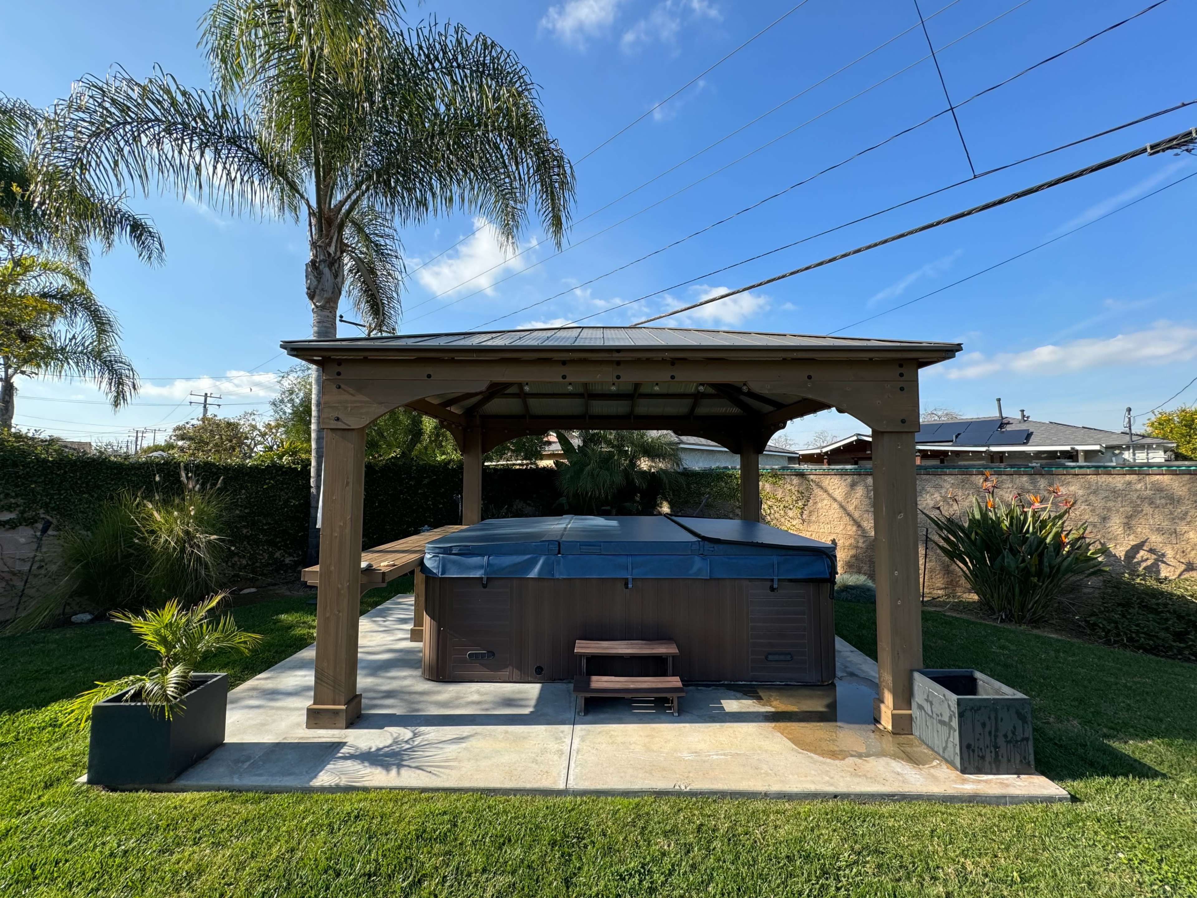 A covered patio with a hot tub is surrounded by grass and palm trees.