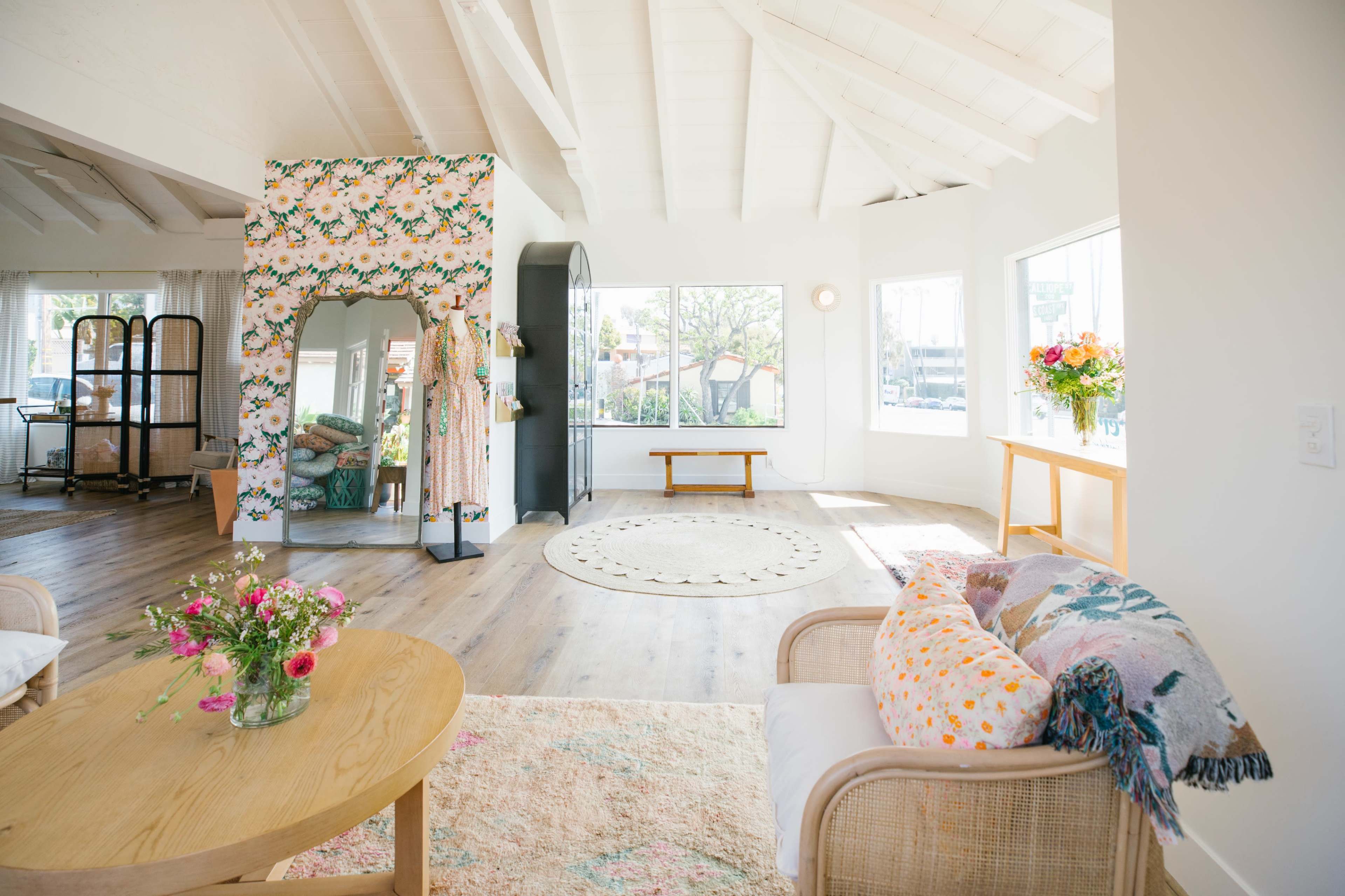 The room features a bright, open space with wooden flooring, large windows, floral decor, and cozy seating.