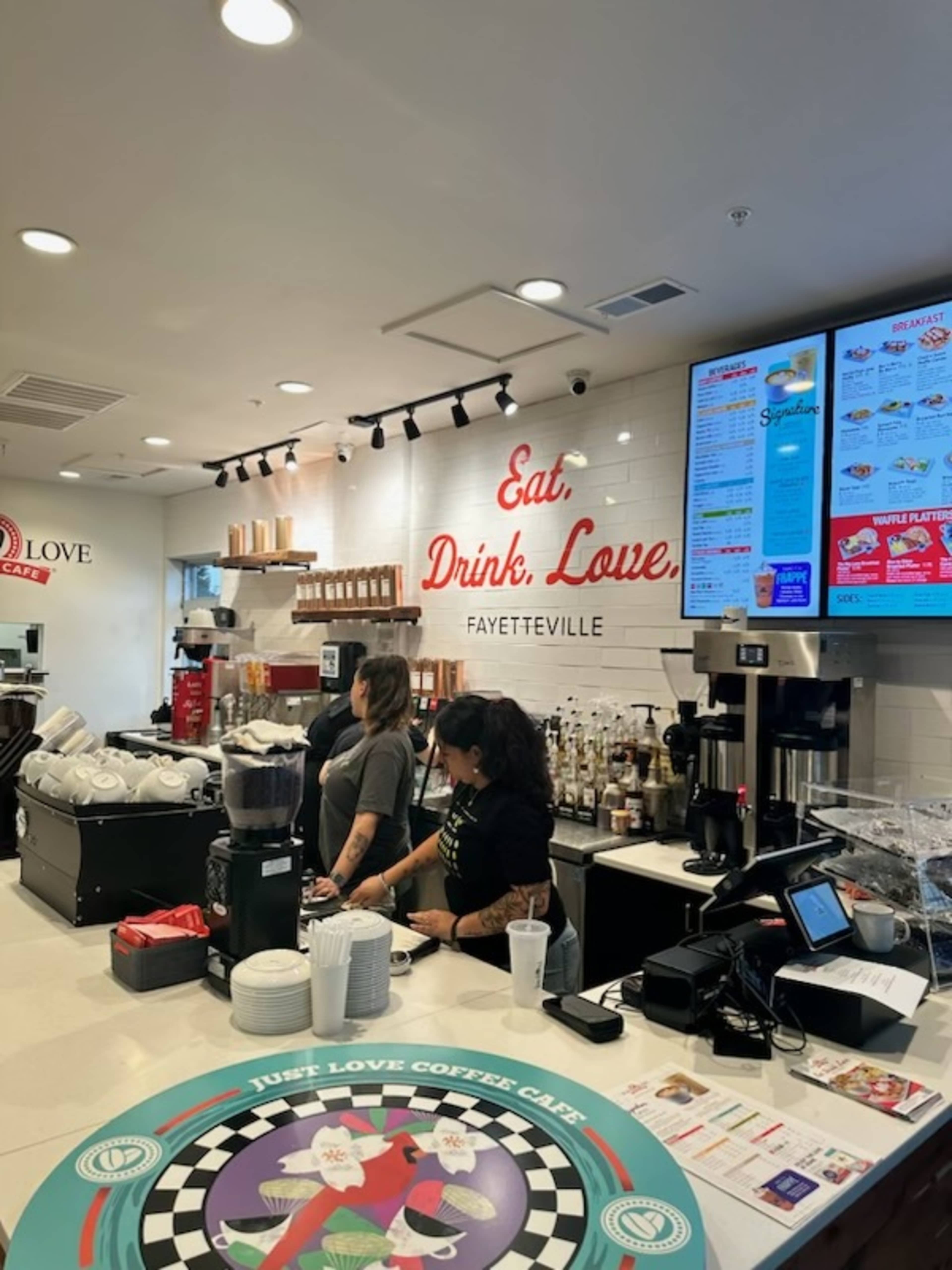 A bustling coffee shop counter features baristas preparing drinks, with a menu displayed on a wall and various coffee supplies on the shelves.