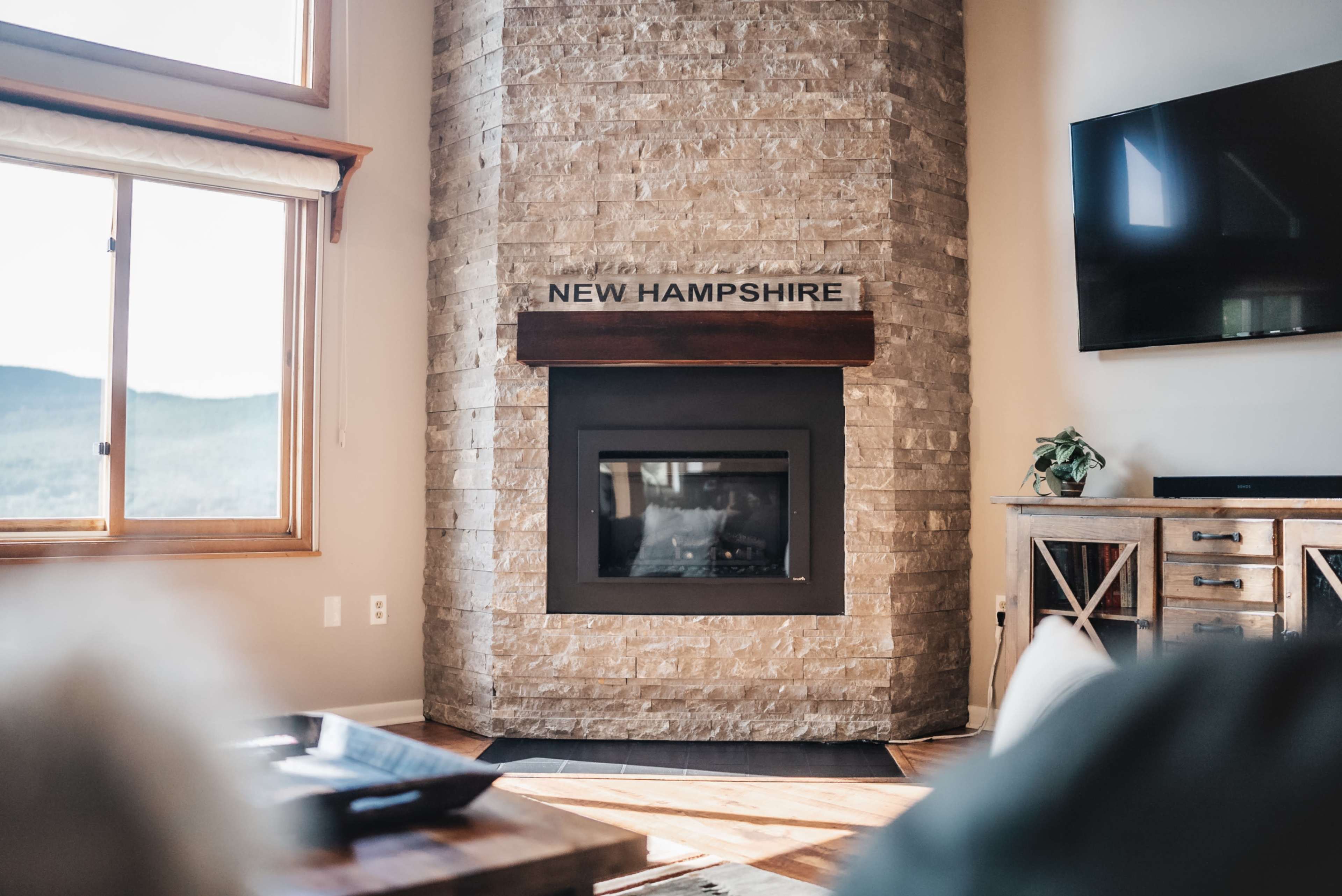 A modern living room features a stone fireplace with a wooden mantel labeled "NEW HAMPSHIRE" and large windows showing a view of the mountains.