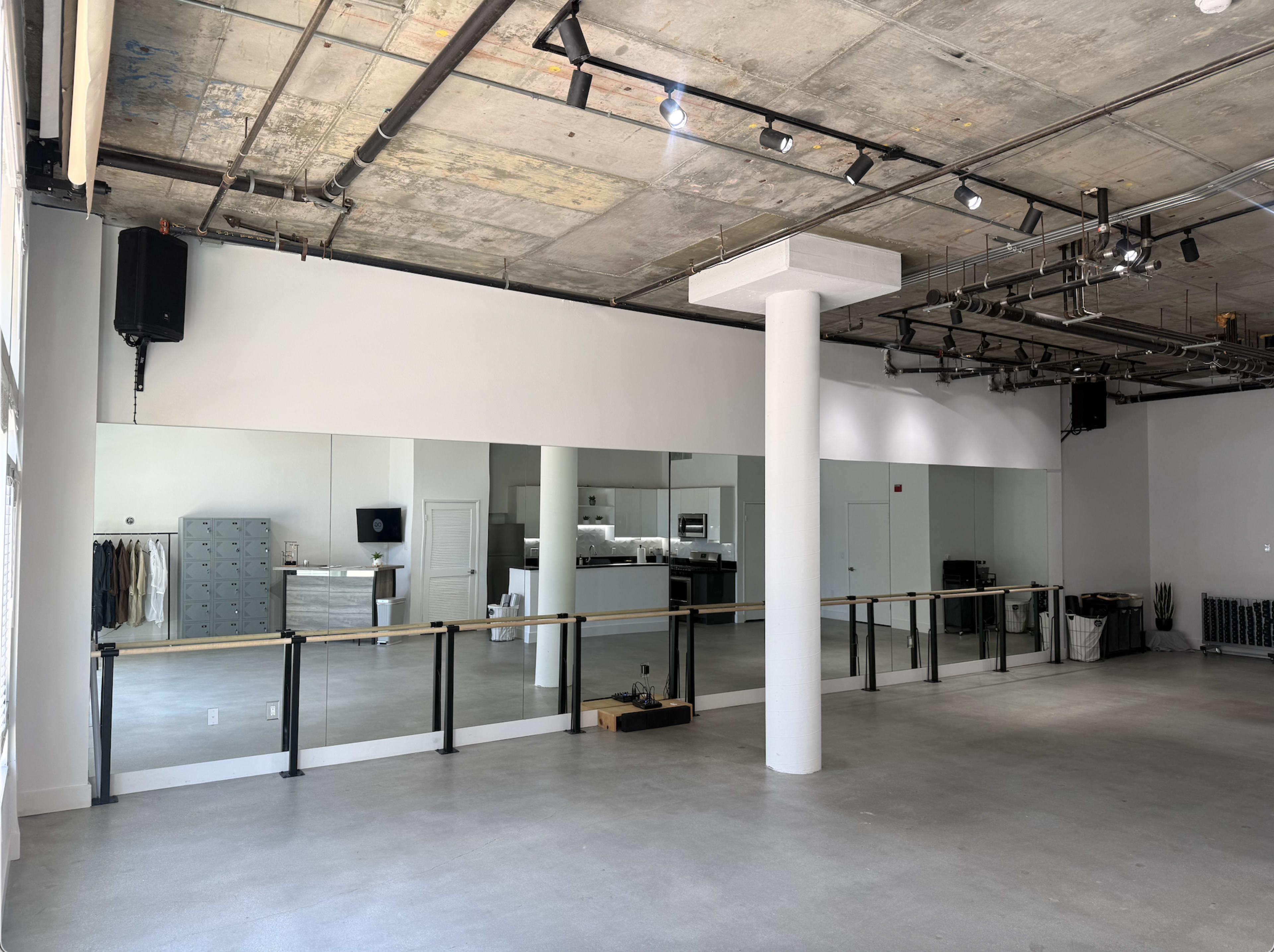 The image shows a spacious, empty studio with tall mirrors on one wall, concrete flooring, and exposed ceiling structures.