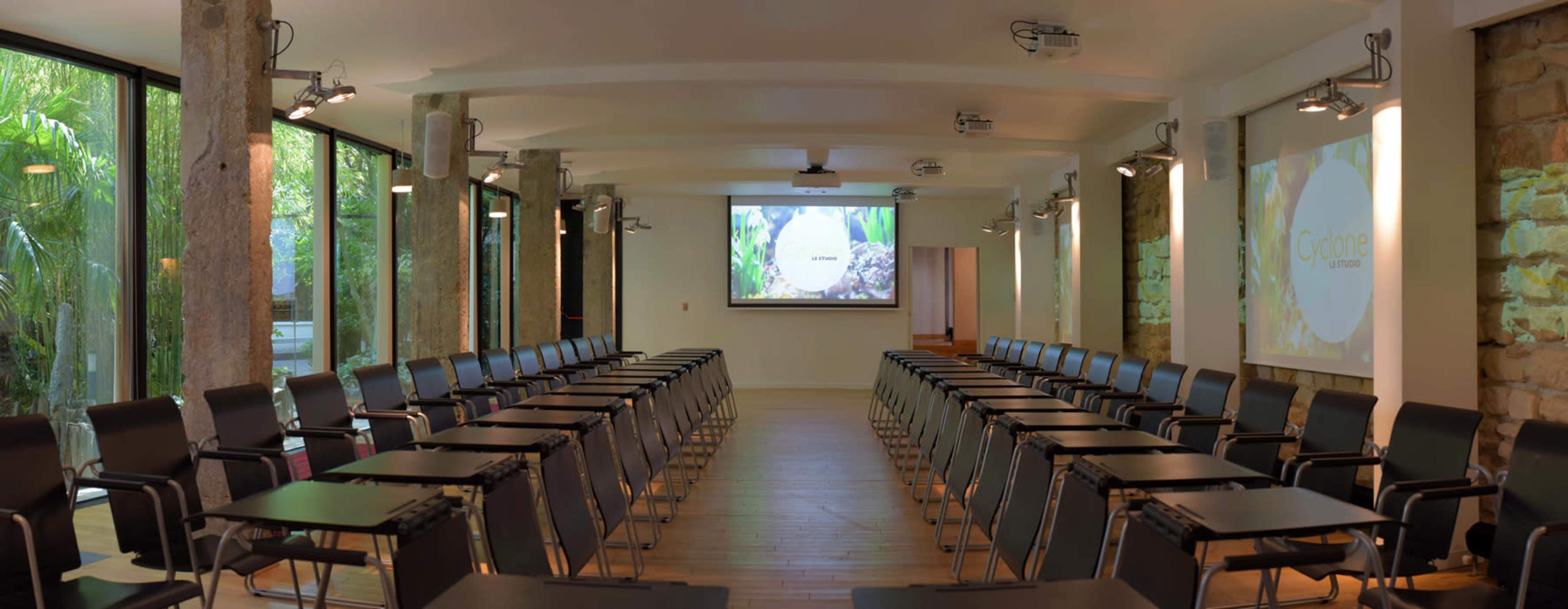 The image shows a conference room with rows of black chairs and small tables facing two projection screens, surrounded by large windows that let in natural light.