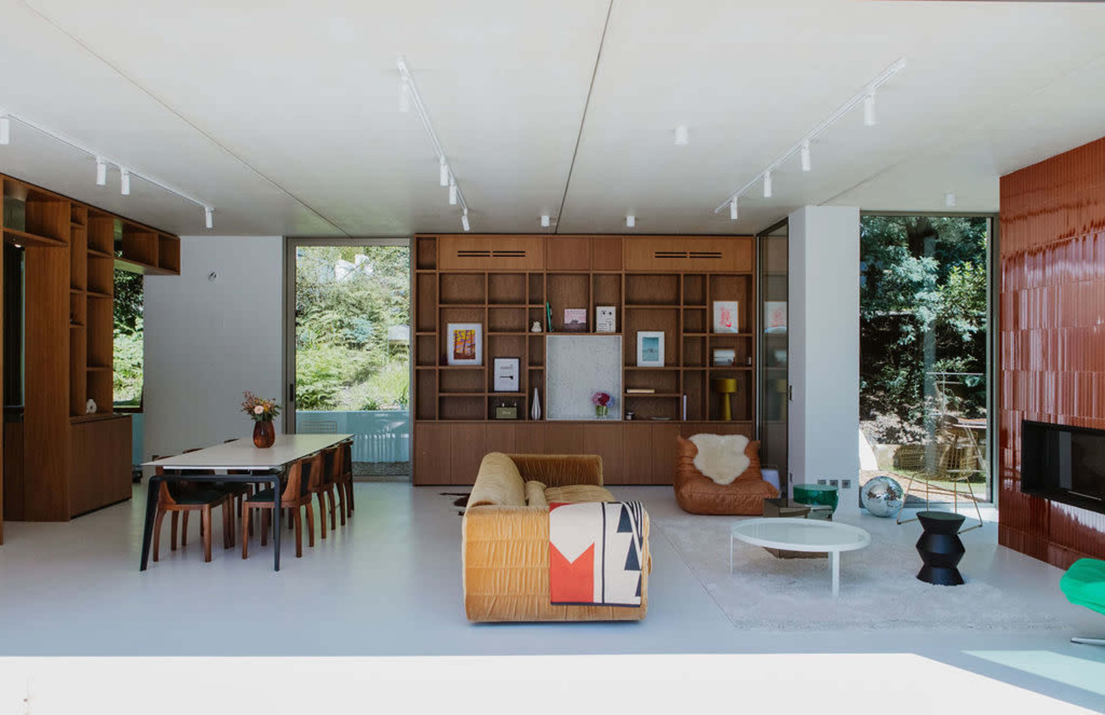 The image features a modern living room with a sofa, a round coffee table, a dining area, and large windows showcasing greenery outside.