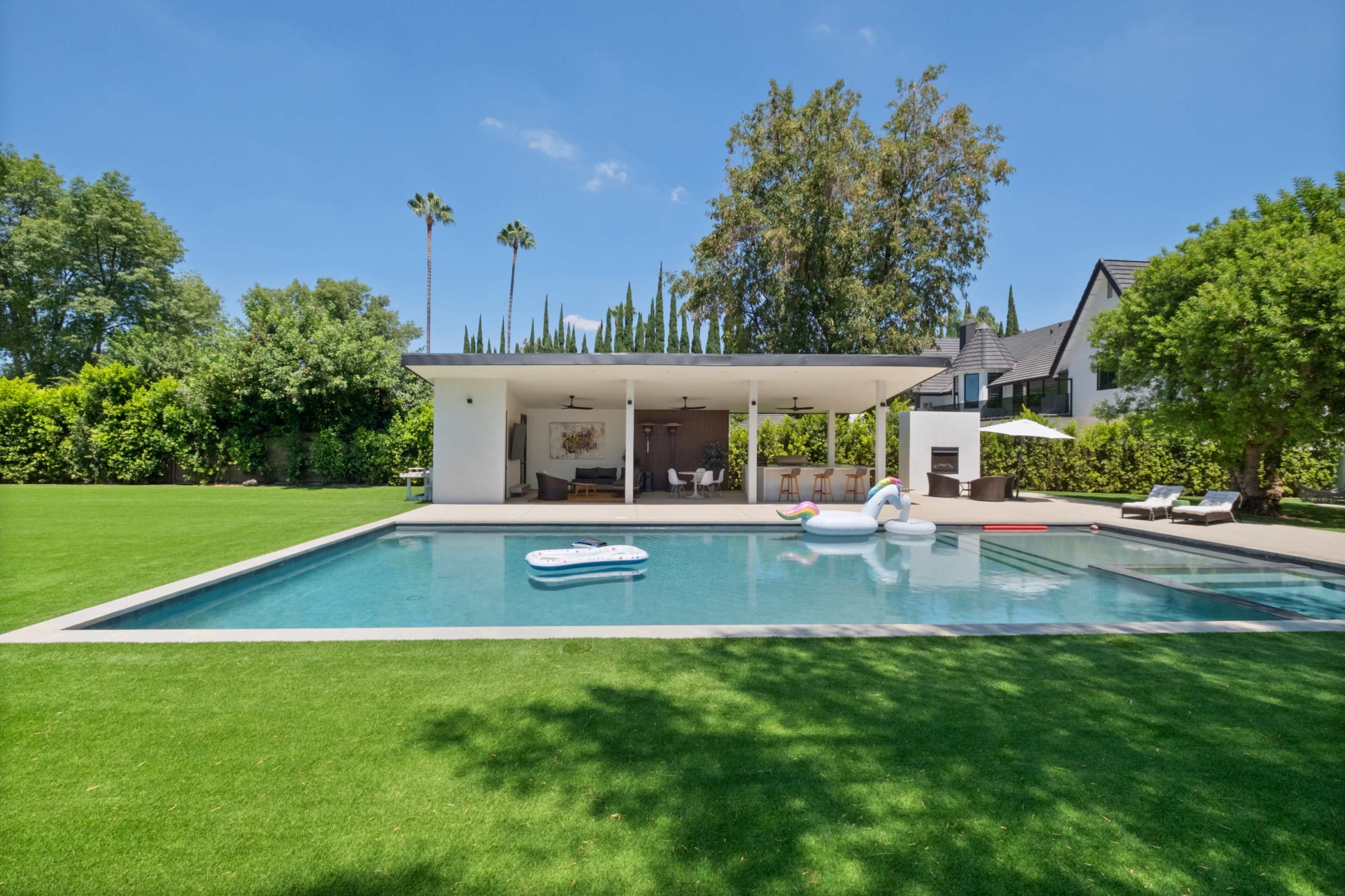 The image shows a modern home with a swimming pool in the backyard, featuring manicured lawns and several lounge chairs.