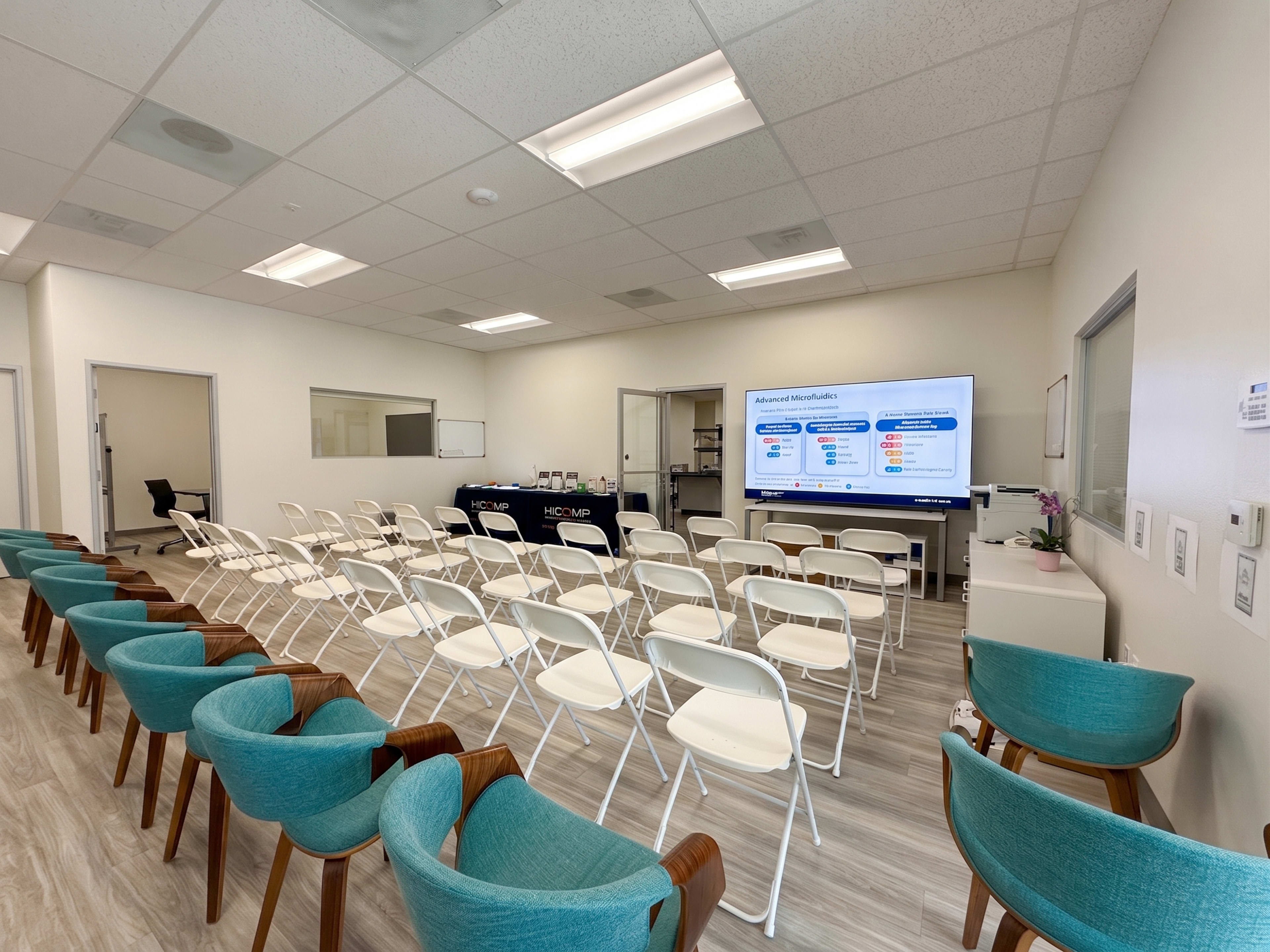 The image shows a waiting room with rows of white chairs and teal armchairs, featuring a large screen displaying information at the front.