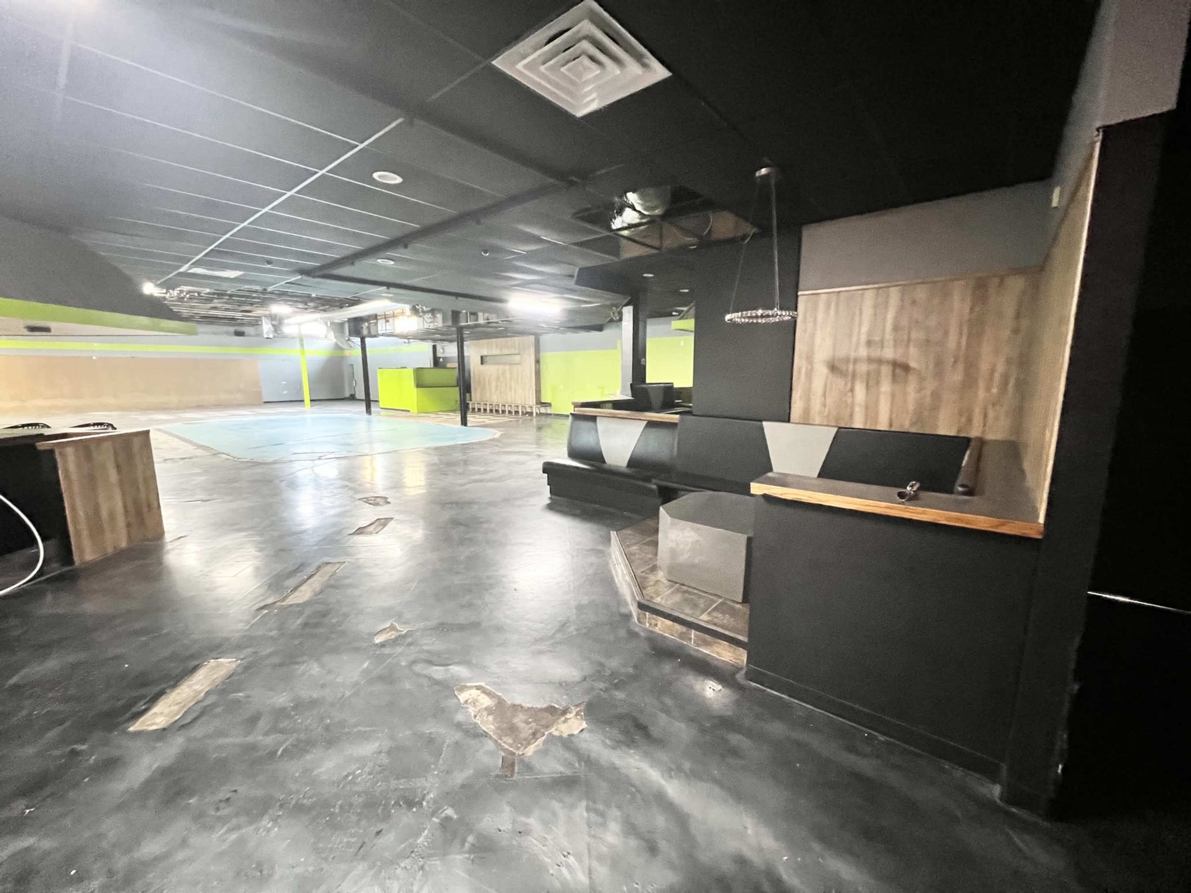 The image shows an empty interior space with a black floor, wooden accents, and a partially visible bar area, illuminated by overhead lights.