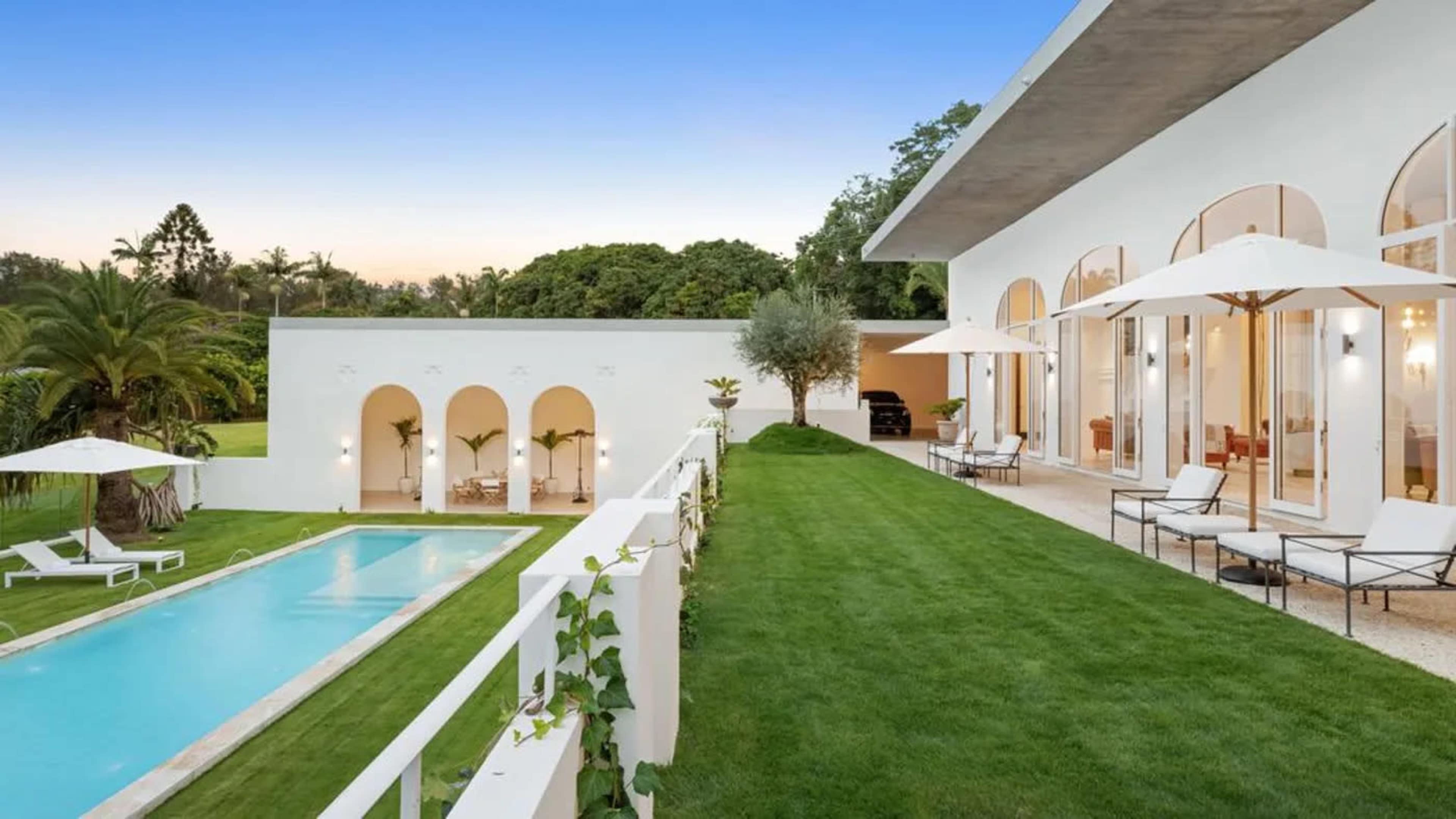 The image shows a modern villa with large windows overlooking a manicured lawn, a swimming pool, and palm trees in the background.