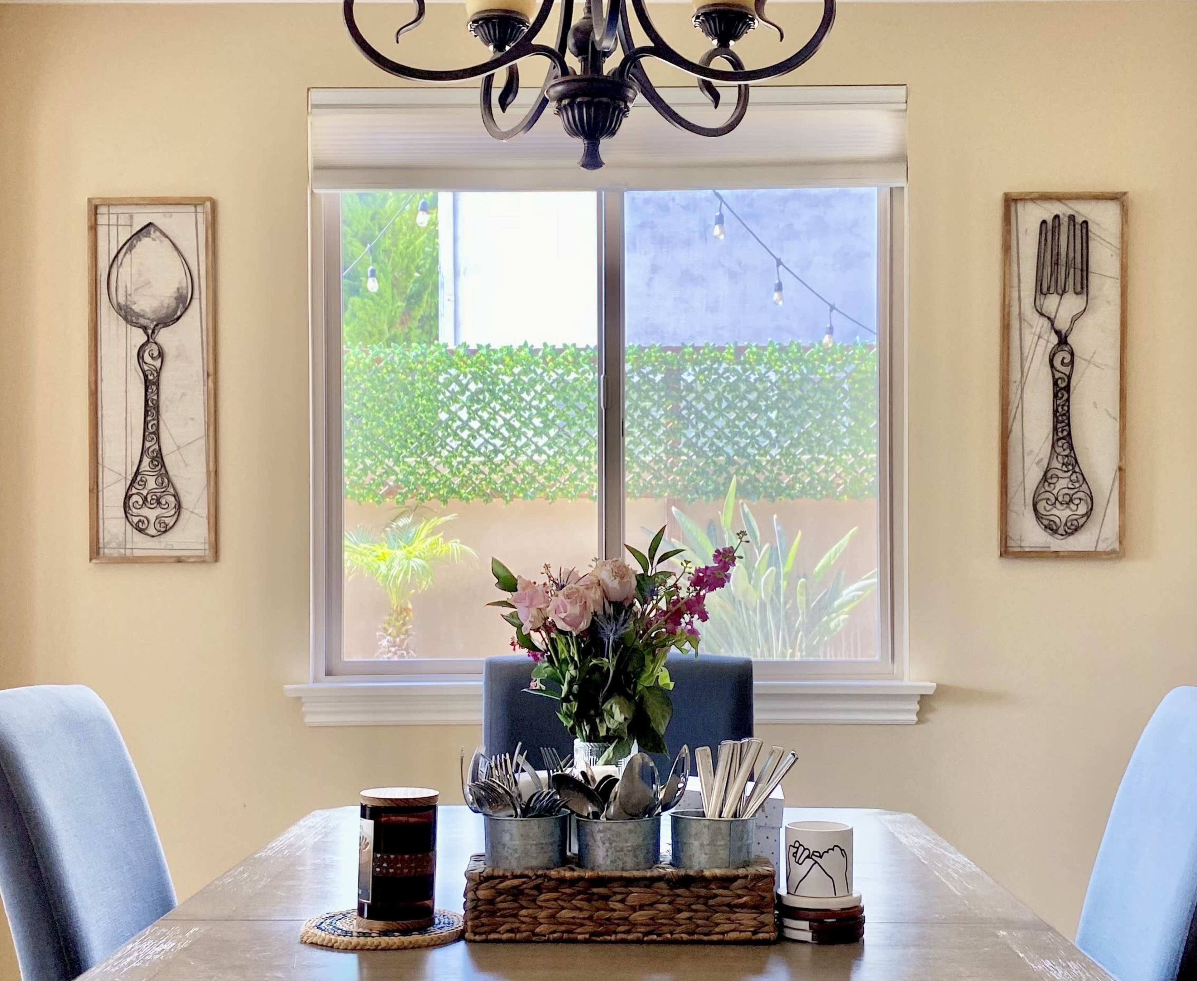 A dining table is set with a flower arrangement and utensils on a tray, flanked by artwork depicting a spoon and fork on the walls, with a view of a garden outside the window.