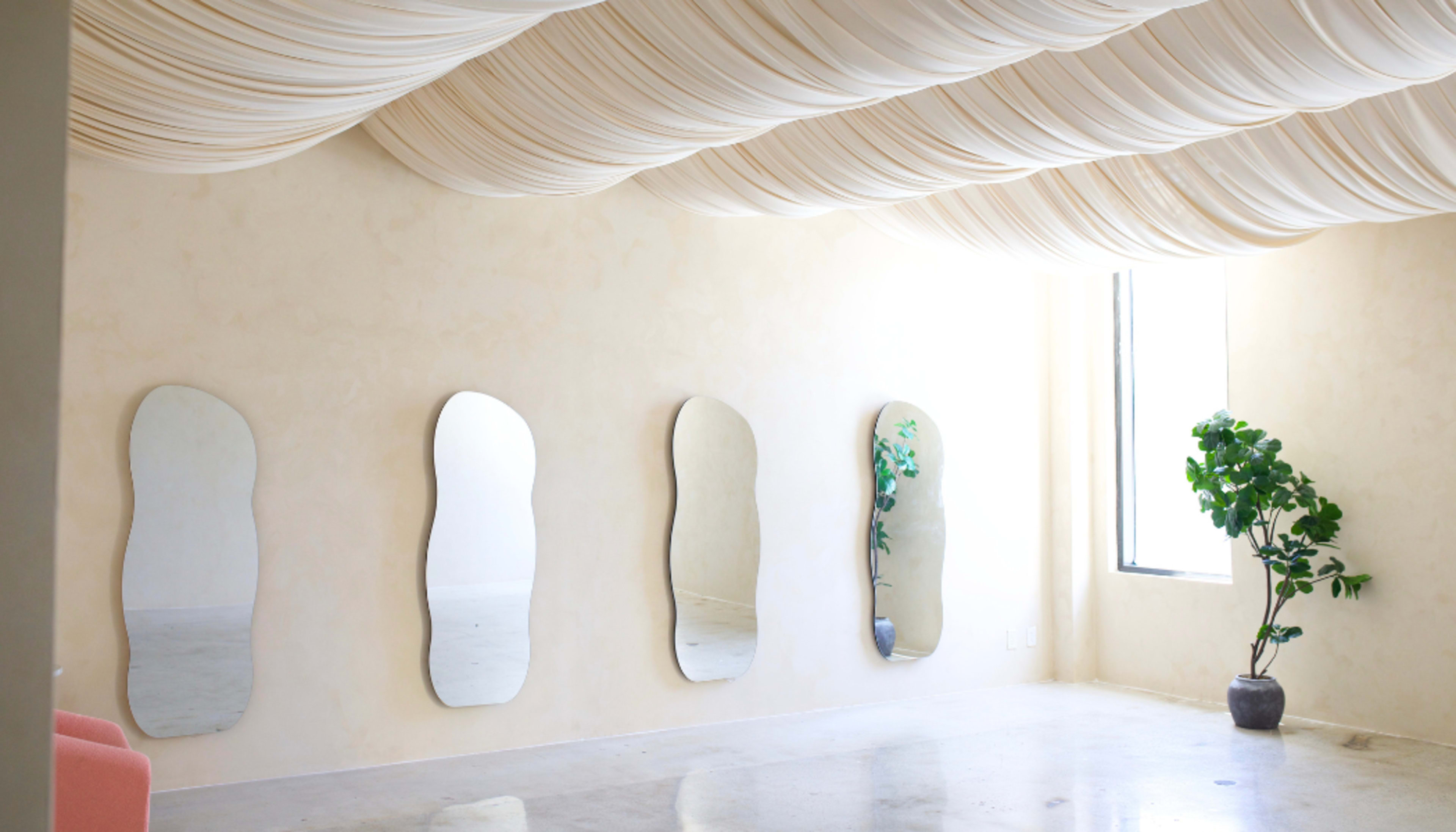 The image shows a spacious room with softly draped fabric ceiling, four elongated mirrors on a light-colored wall, and a potted plant near a window.