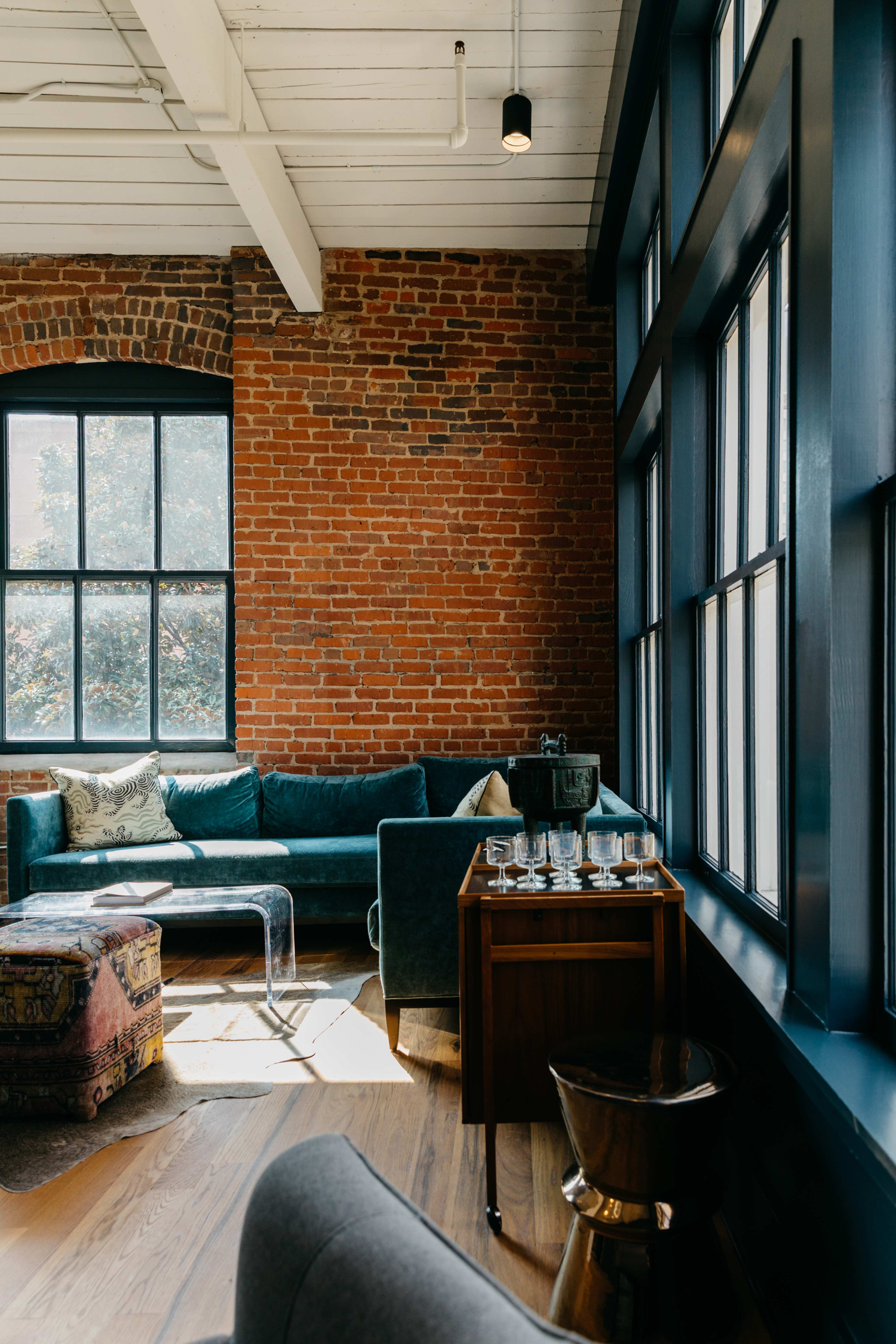 A cozy living space featuring a brick wall, a teal sofa, a wooden side table with glasses, and large windows letting in natural light.