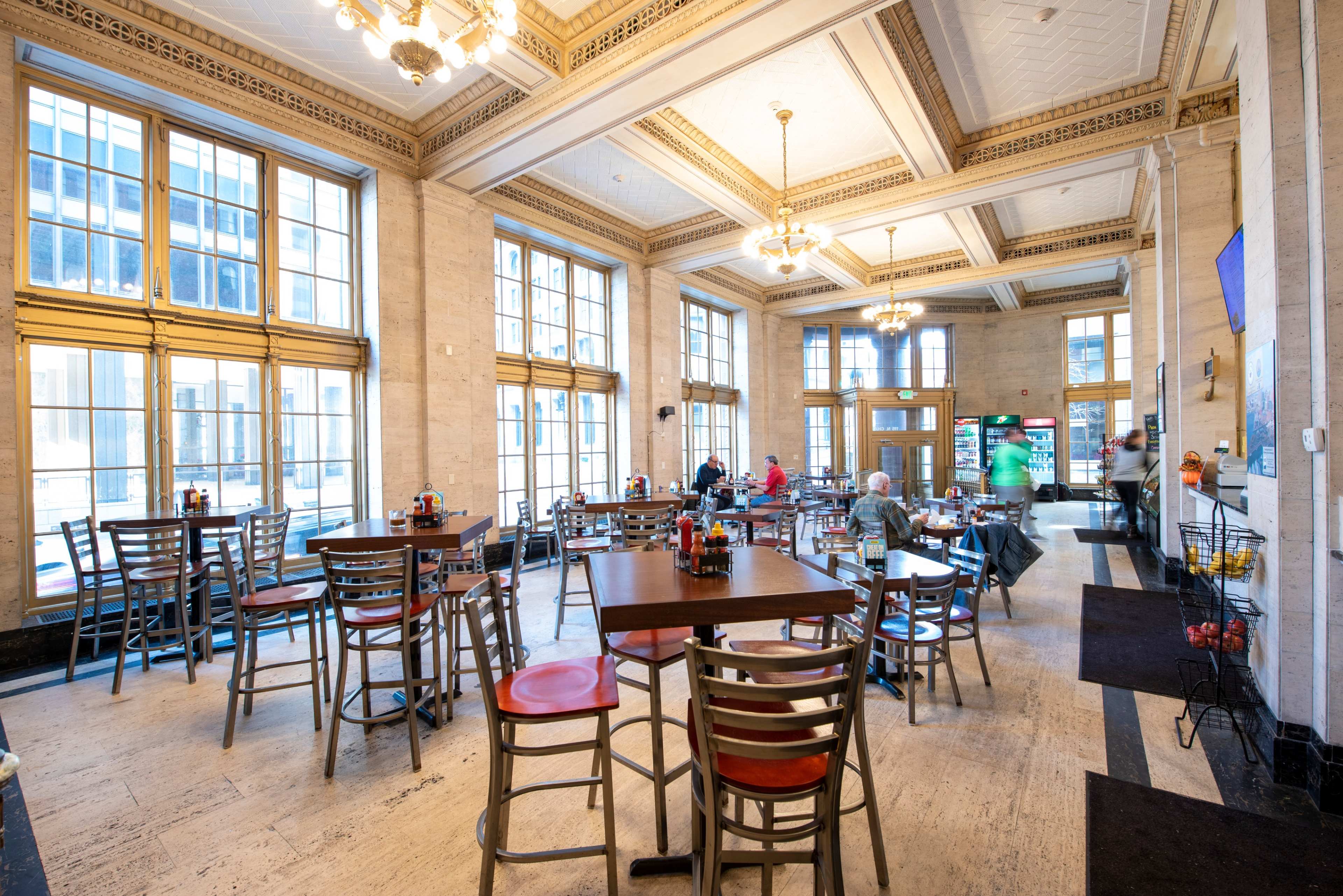 A spacious café area with tables and chairs, large windows letting in natural light, and patrons seated at various tables.