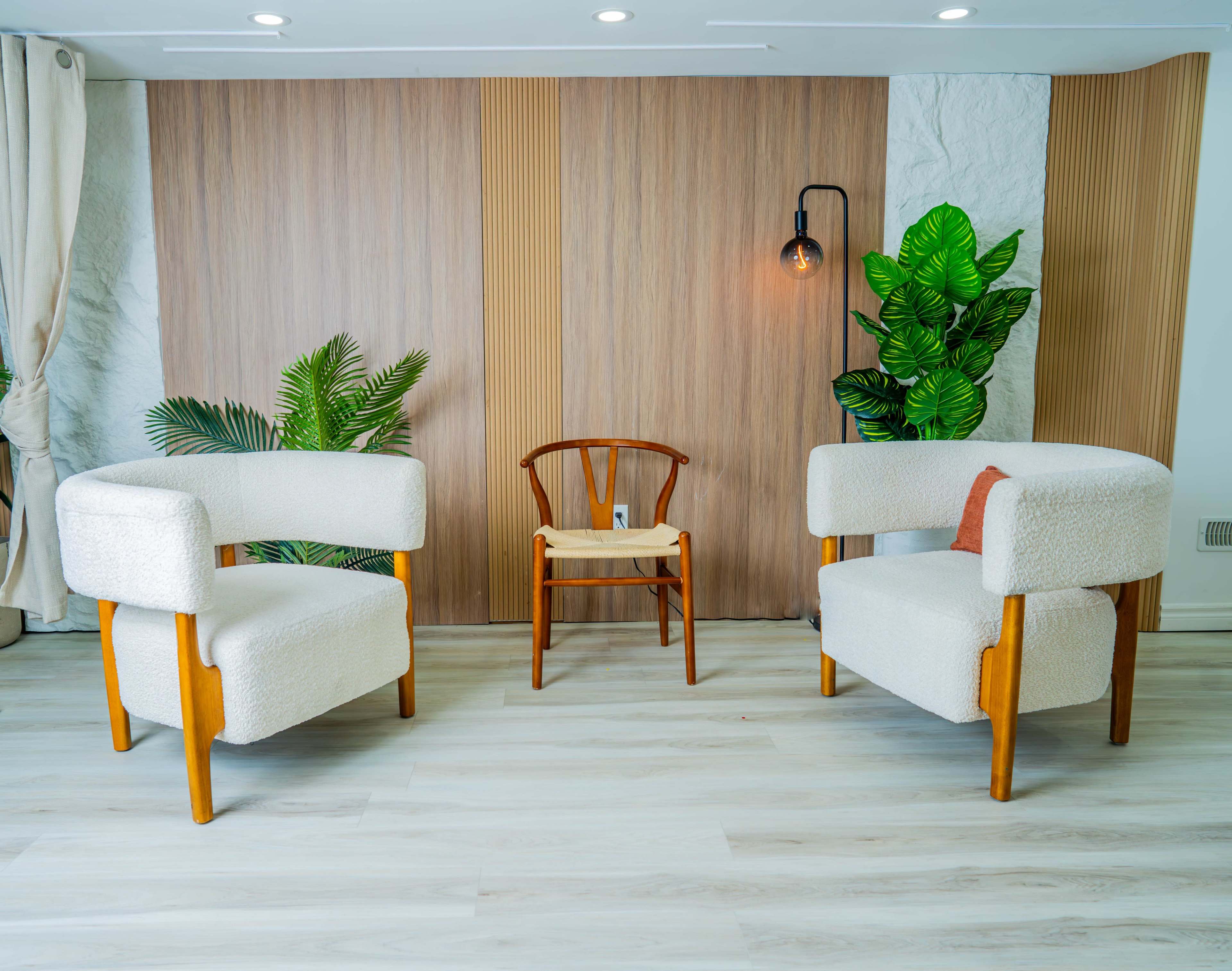 The image shows a minimalist seating arrangement with two upholstered chairs on either side of a wooden chair against a backdrop of wooden panels and decorative plants.