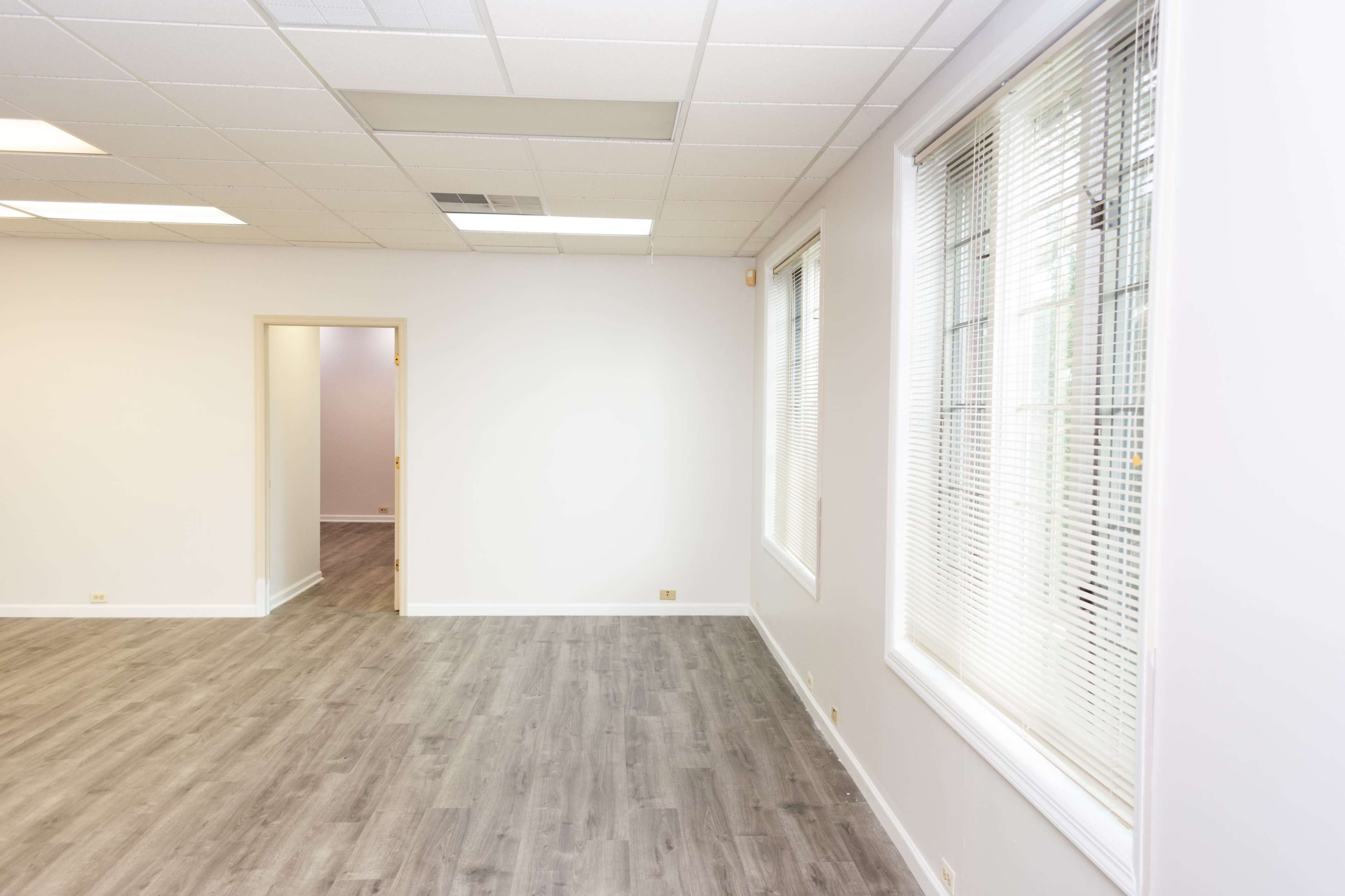 The image shows a spacious, unfurnished room with gray flooring, large windows, and a doorway leading to another room.