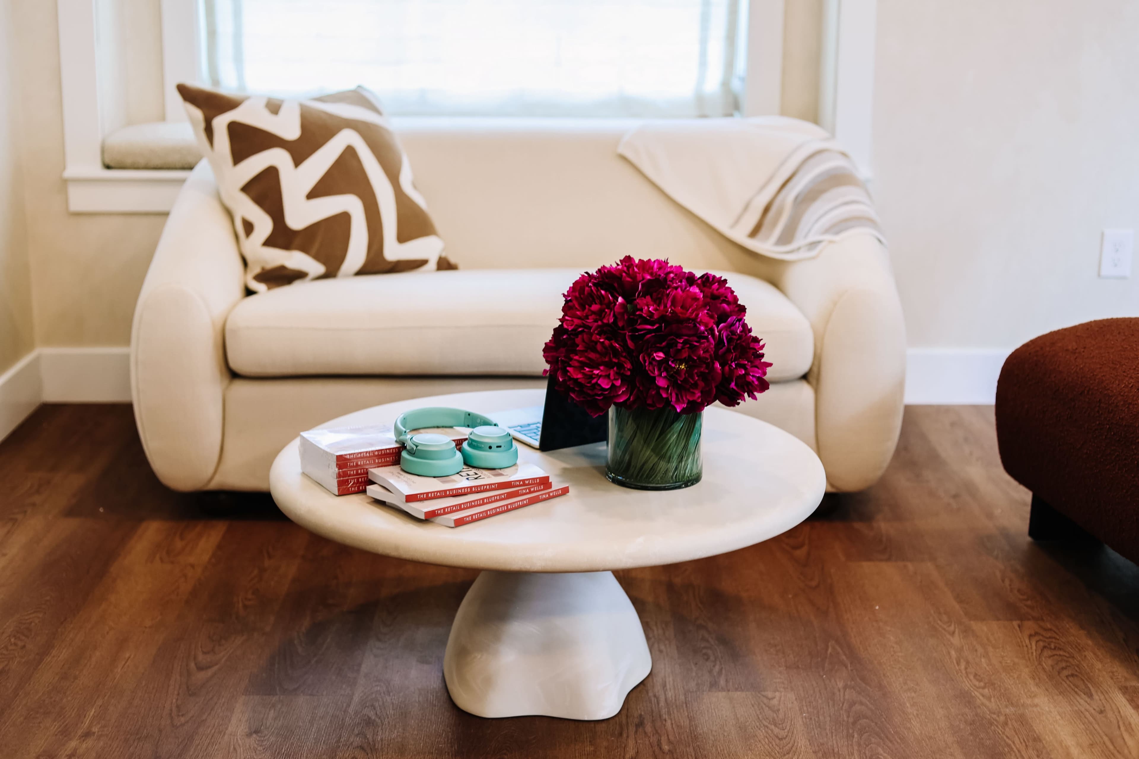A coffee table with a marble top displays books and decorative items, including a turquoise bowl and a vase of red flowers, in a living room setting featuring a light-colored sofa.