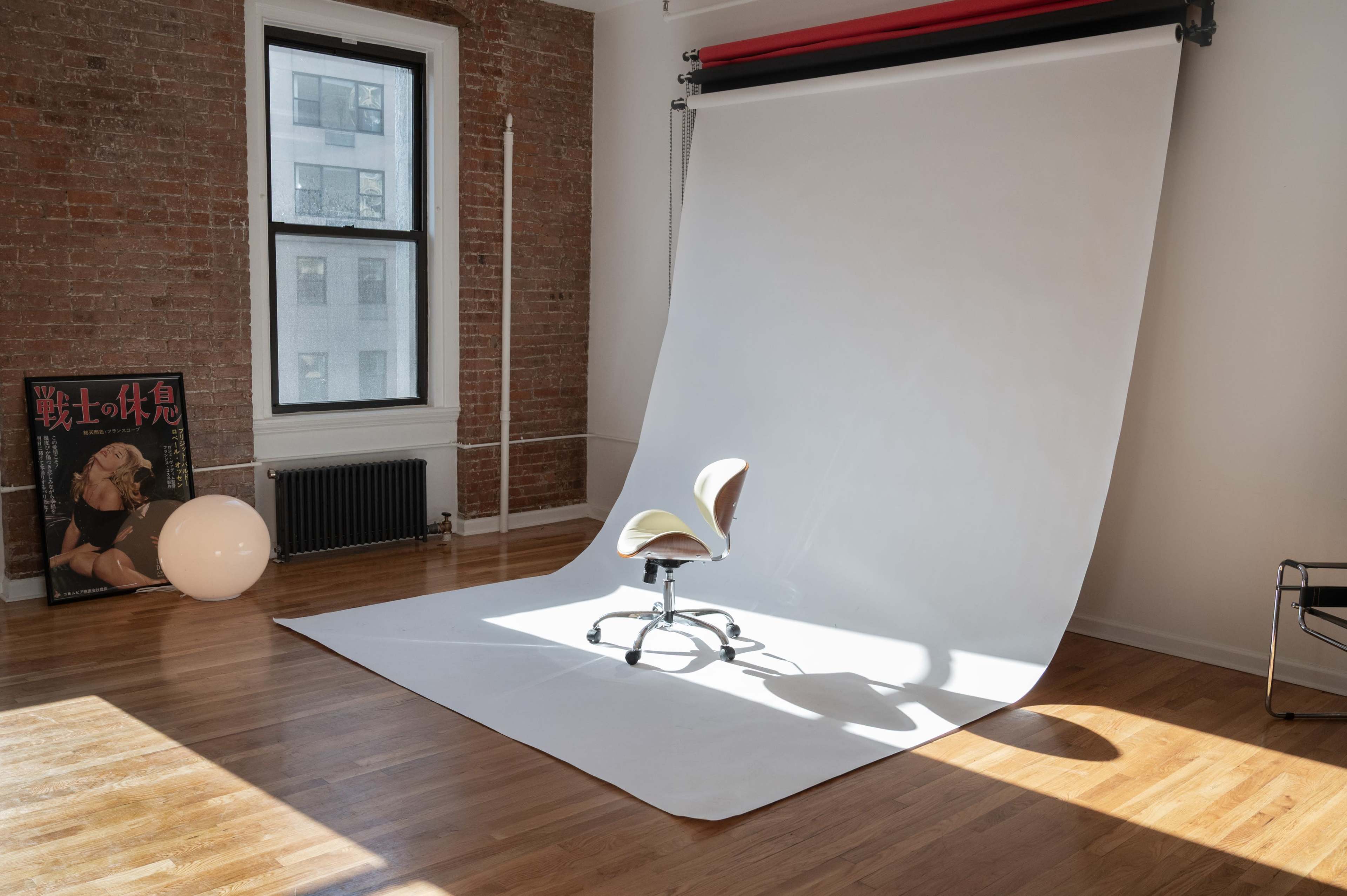 The image shows a photography studio with a white backdrop, an office chair in the center, and a round light fixture beside a framed poster on a brick wall.