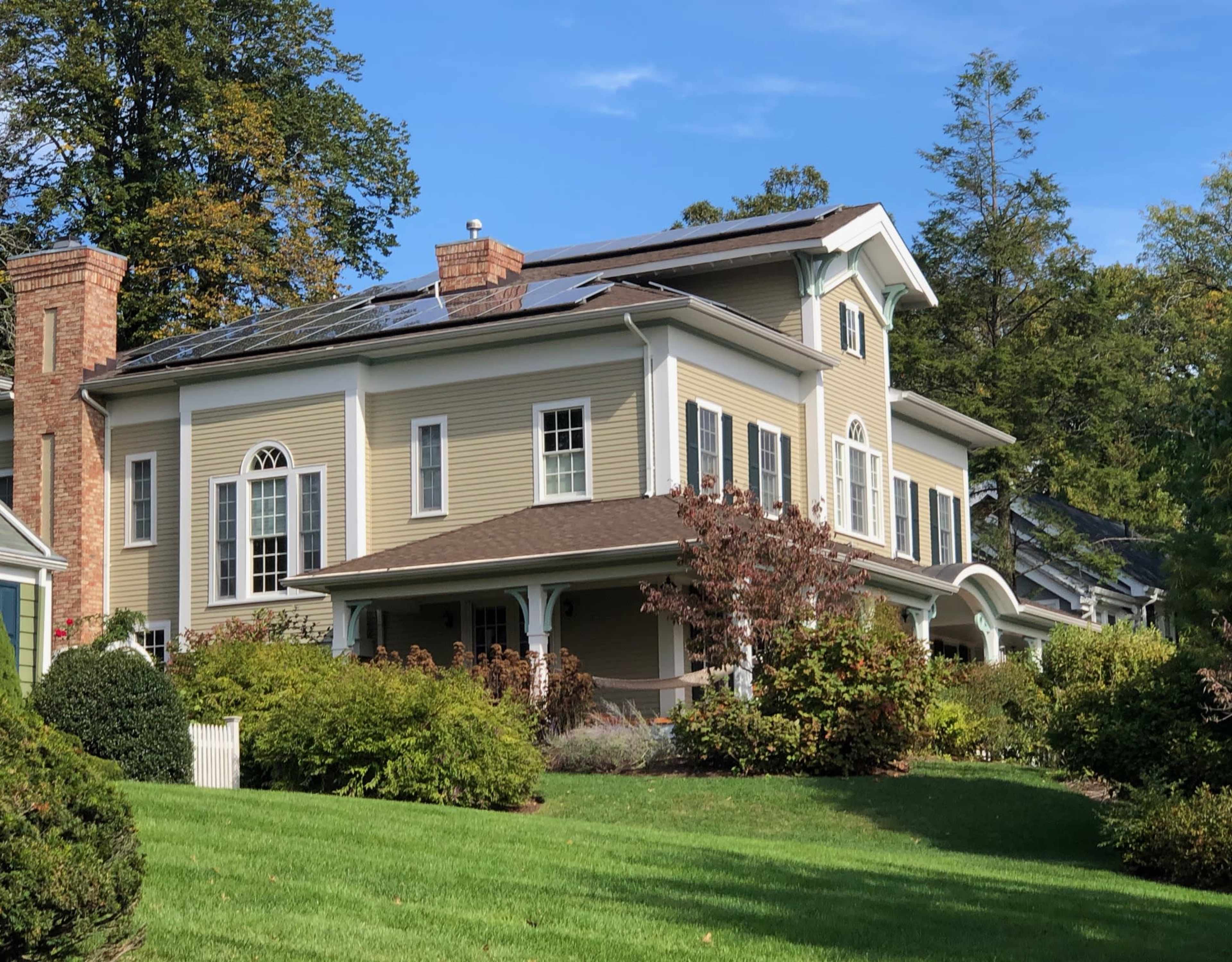 The image shows a large, two-story house with solar panels on the roof, surrounded by a well-manicured lawn and landscaping.