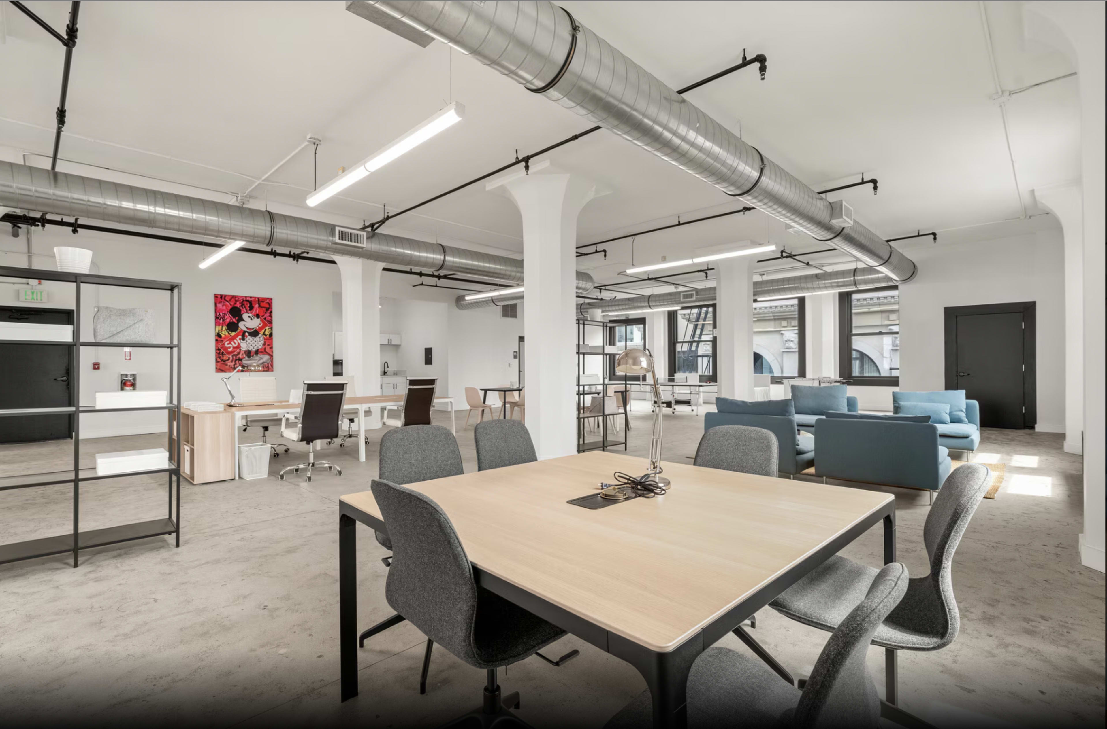 The image shows a modern office space with a large central table, several workstations, and a lounge area featuring a blue sofa.