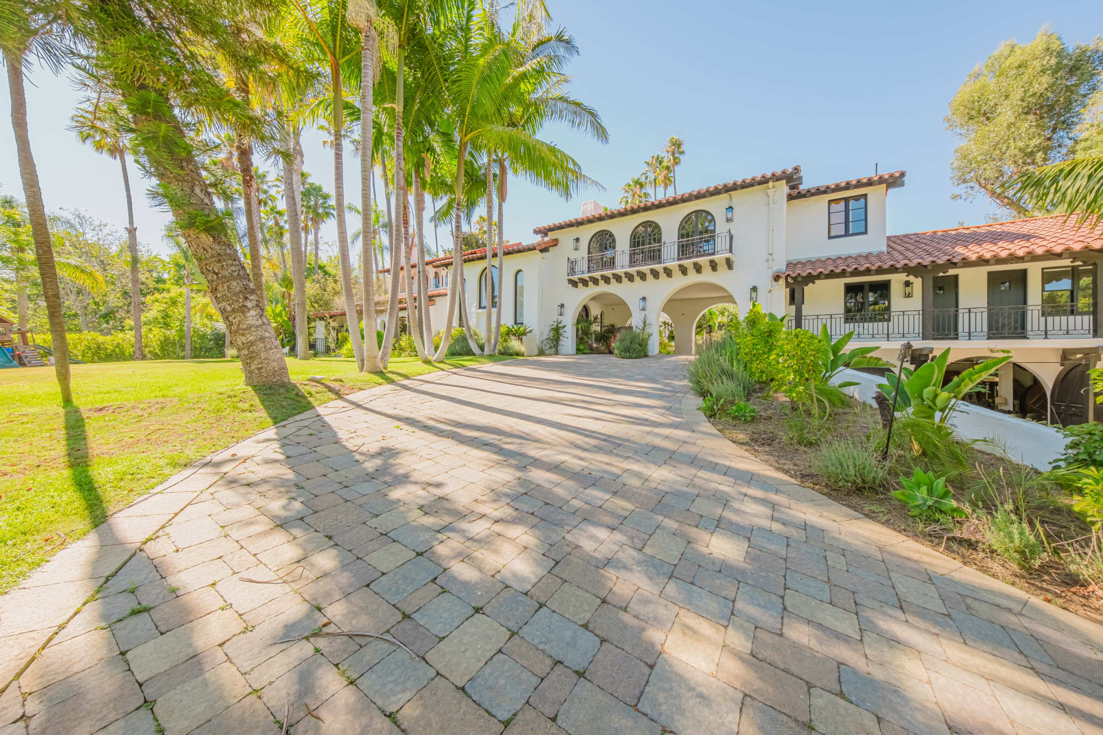 The image shows a spacious, upscale home with a tiled pathway leading to a front entrance, surrounded by palm trees and lush greenery.