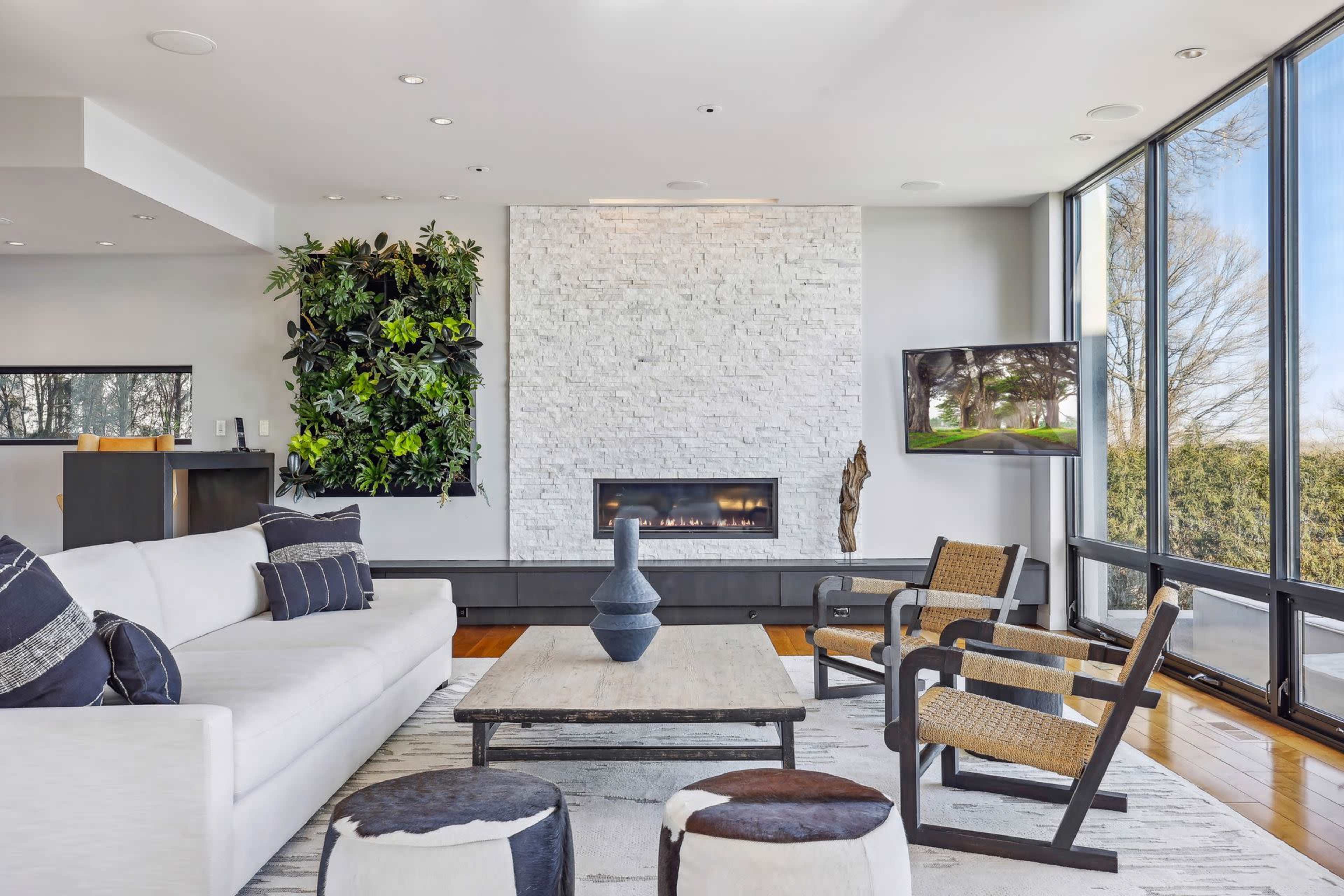 The image shows a modern living room featuring a white sofa, a stone fireplace, a coffee table, and large windows with a view of greenery outside.