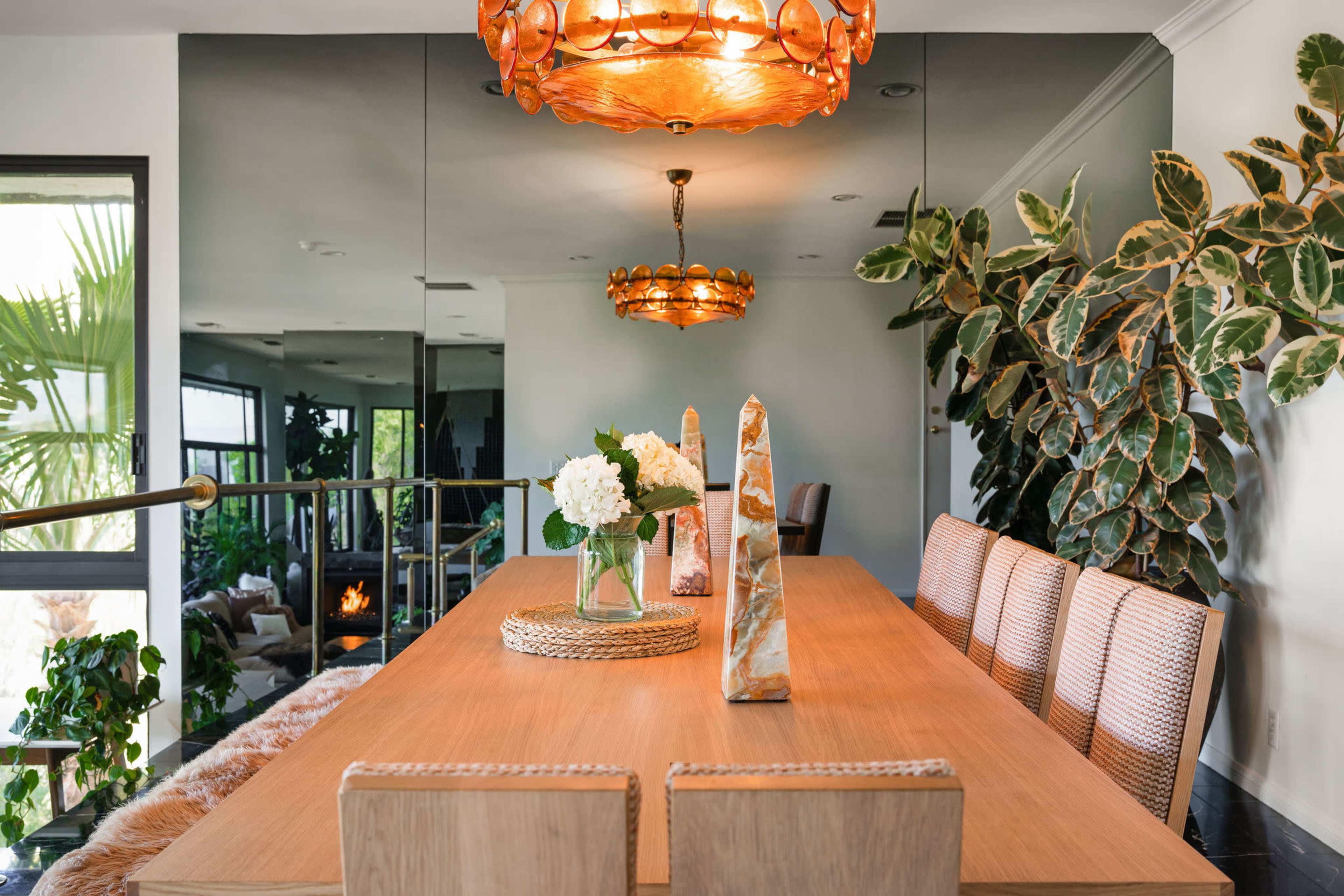 A dining area features a wooden table surrounded by woven chairs, adorned with decorative stone pieces and a vase of flowers, under a decorative lamp and adjacent to indoor plants.