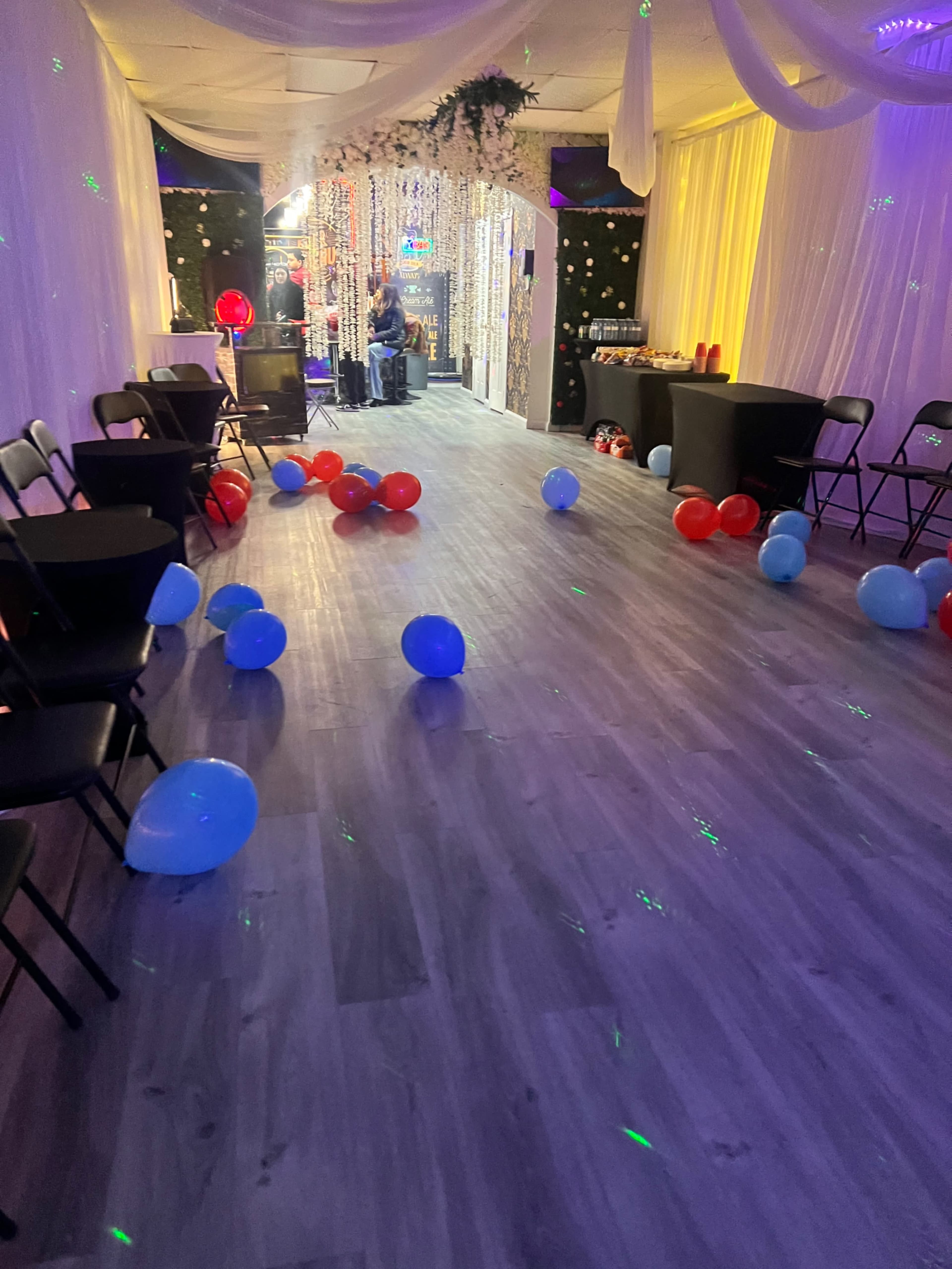 The scene shows a decorated party venue with scattered red and blue balloons on the floor and string lights in the background.