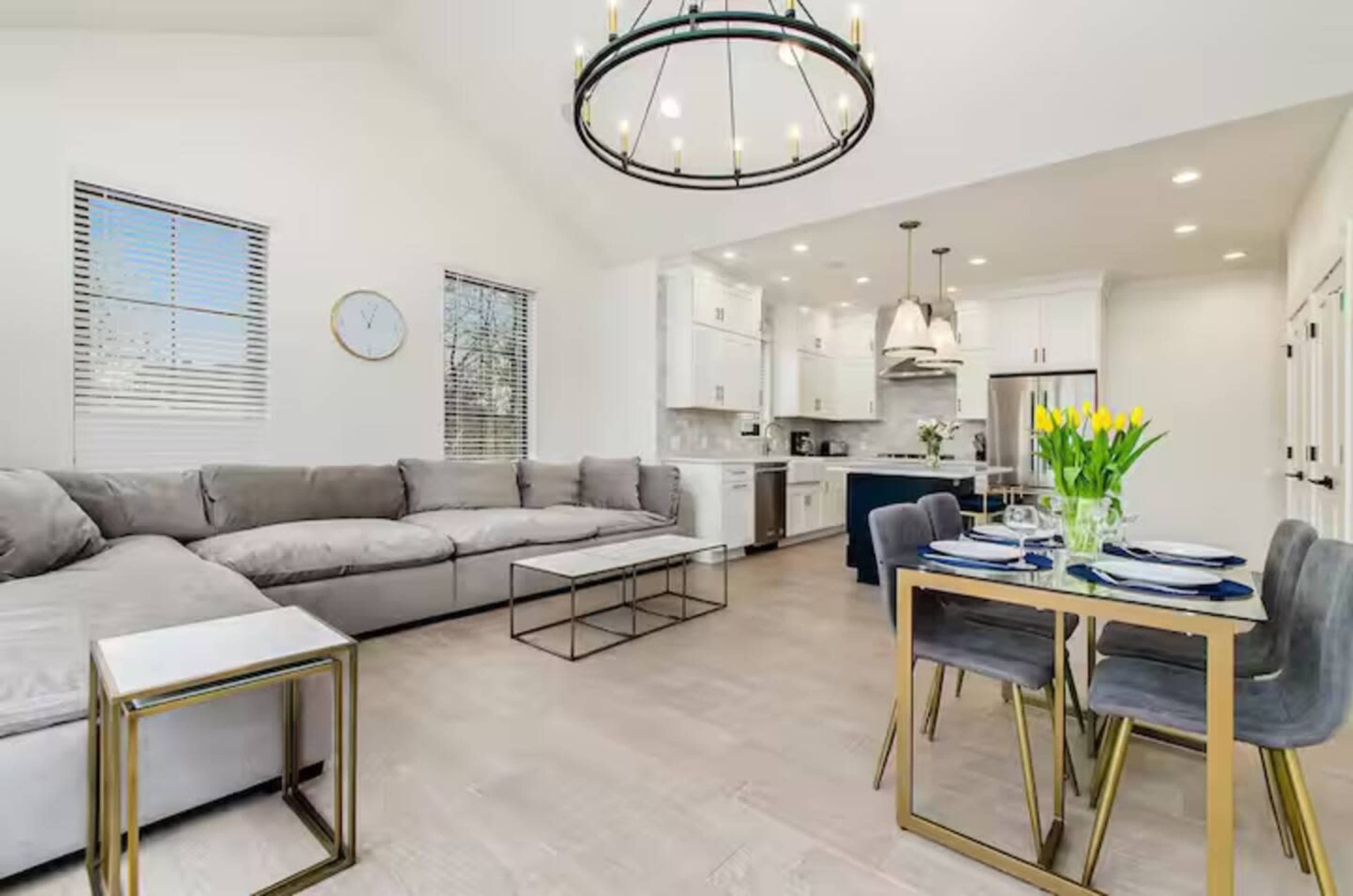The image shows a modern living area with a large sectional sofa, a dining table set for four, and a well-lit kitchen in the background.