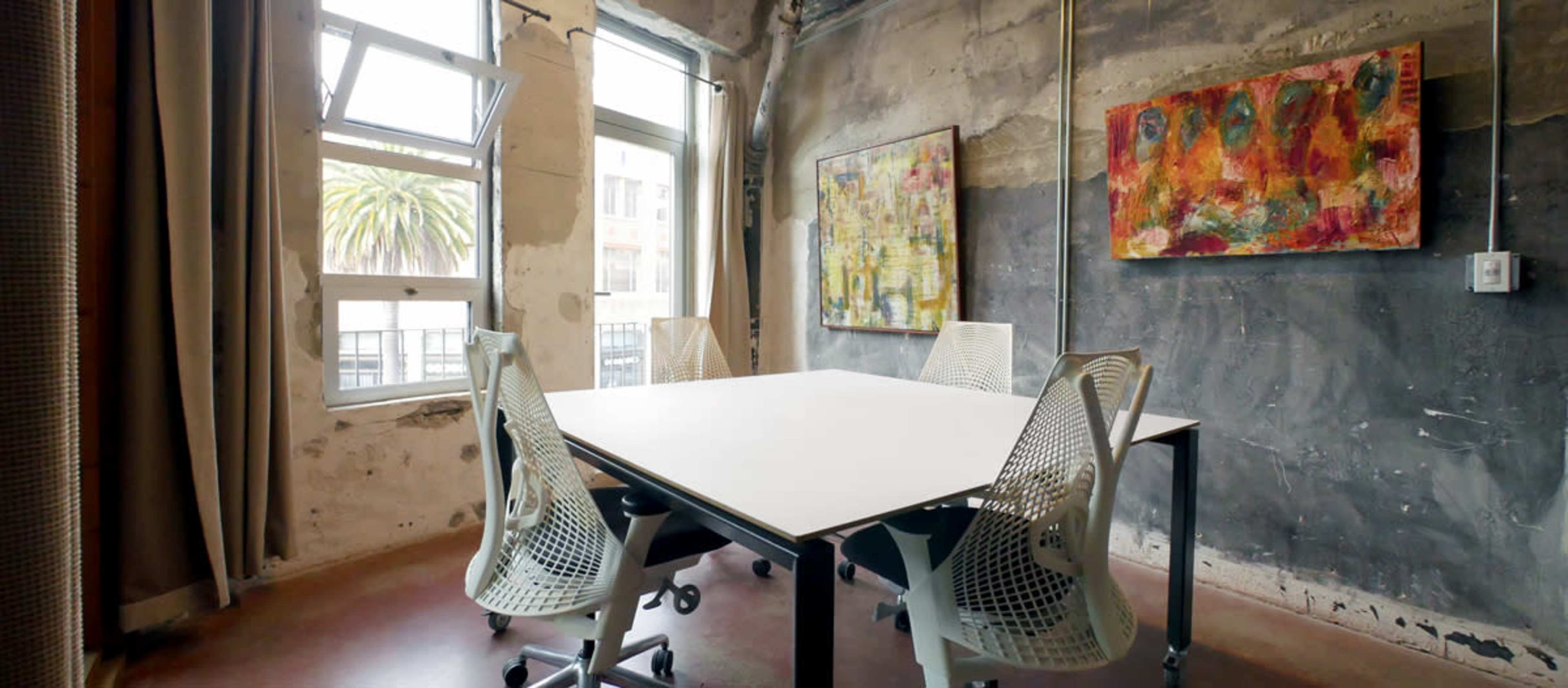 A conference room features a white table surrounded by four ergonomic chairs, with abstract artwork on the walls and large windows letting in natural light.