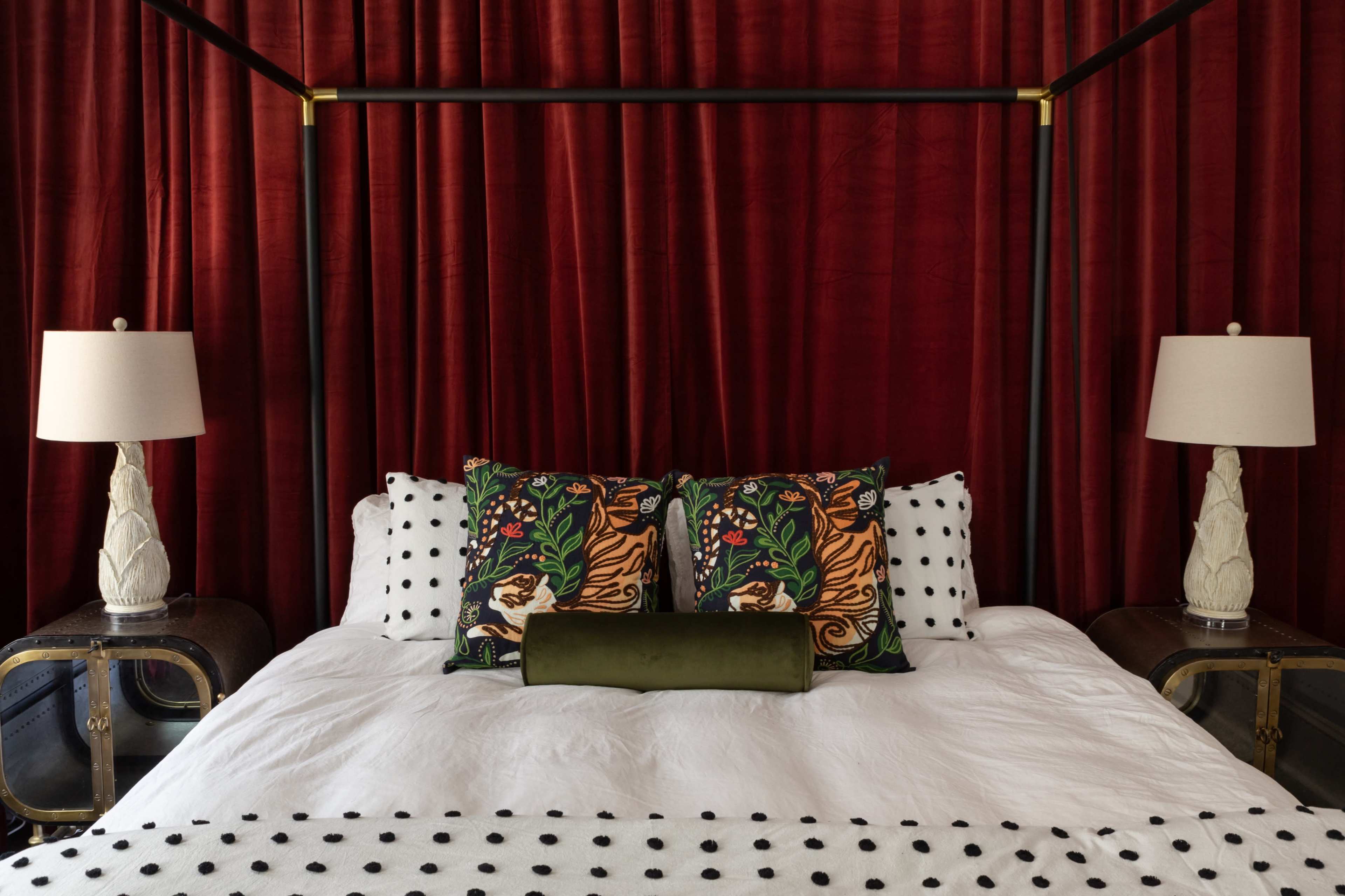 A four-poster bed is centered in a room with red velvet curtains, flanked by two lamps on wooden side tables.