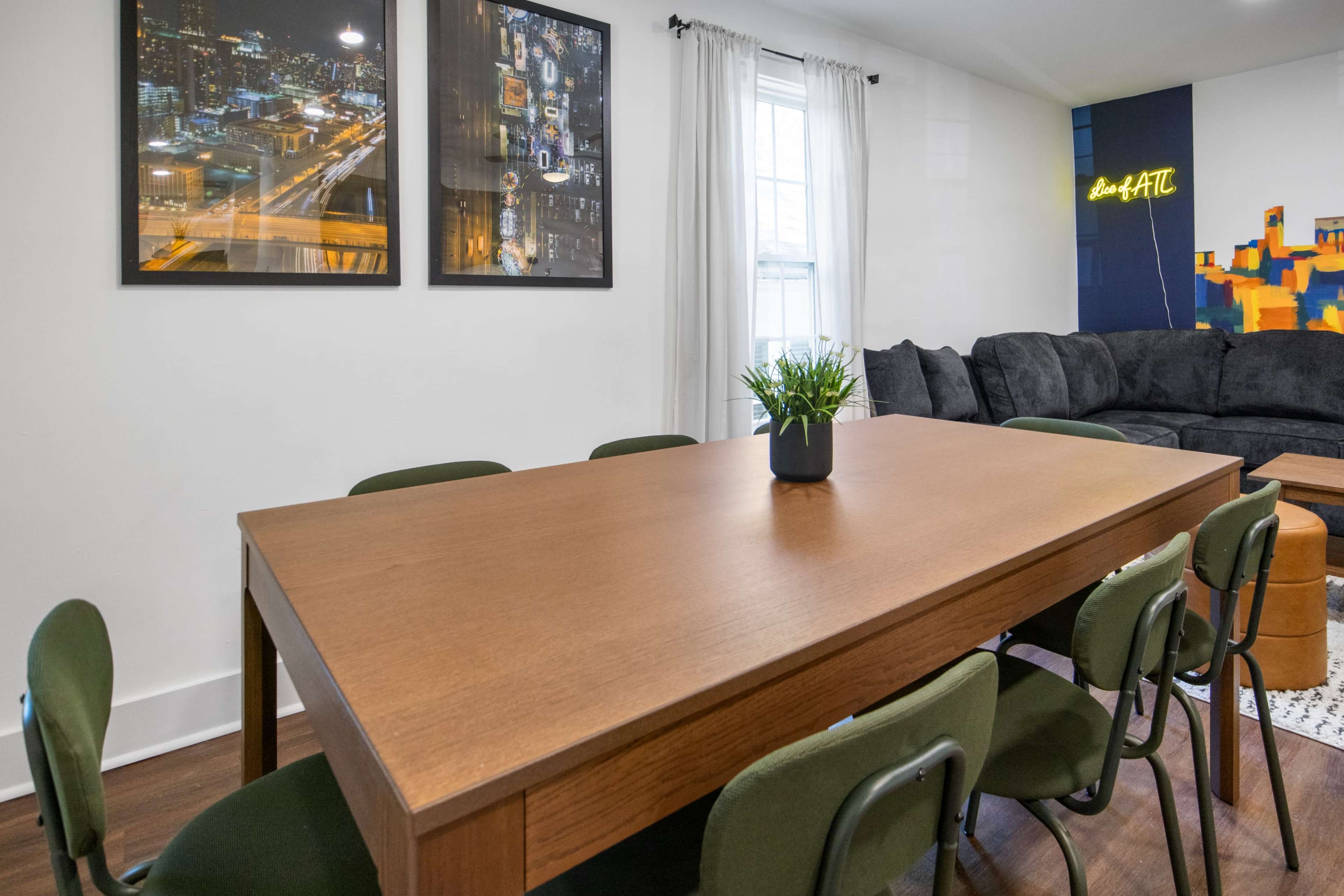 A dining area with a wooden table surrounded by green chairs, set under two framed cityscape photographs and a vibrant wall display.