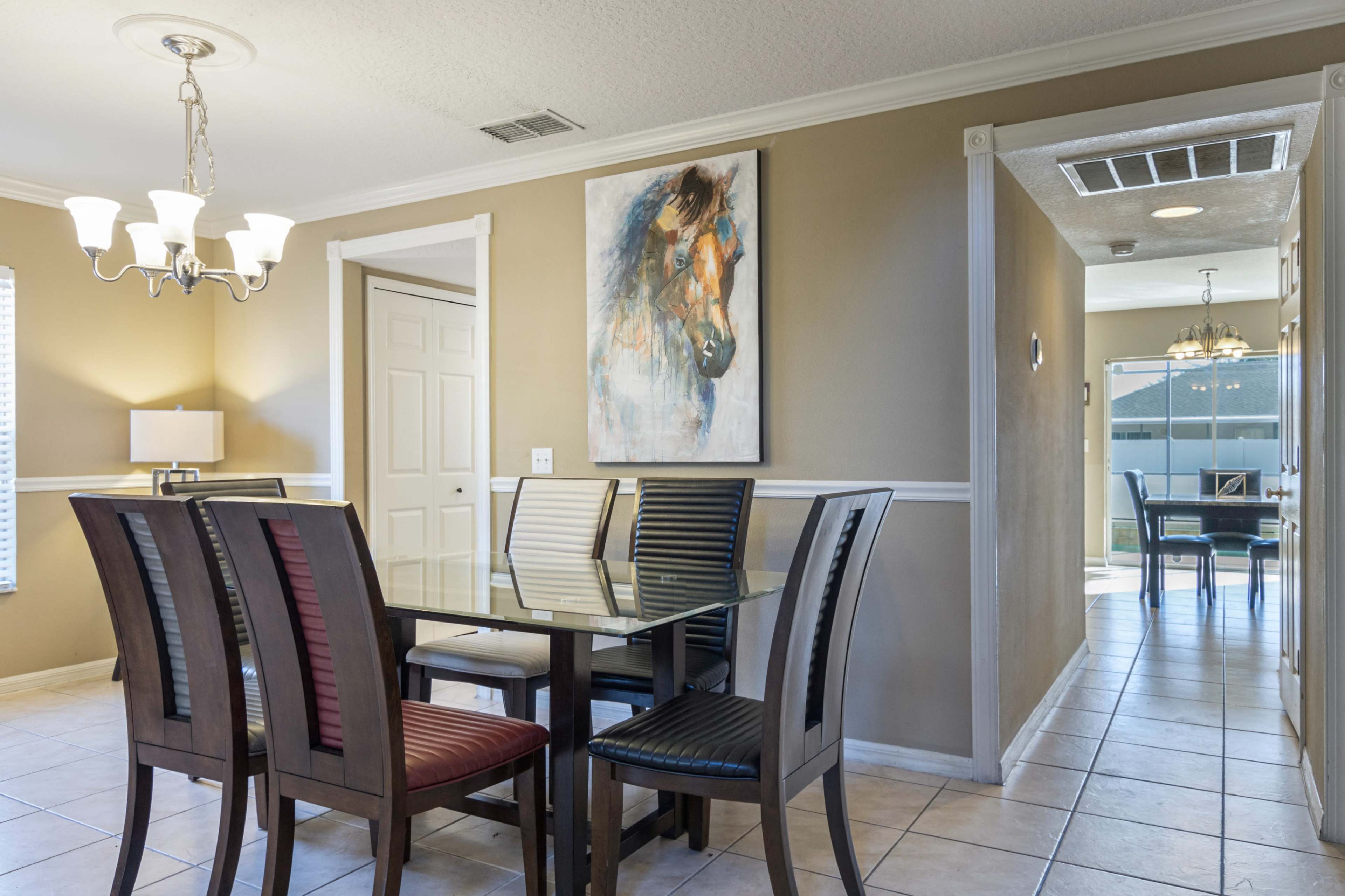 A glass dining table surrounded by wooden chairs is positioned in a well-lit room featuring a painting on the wall and a doorway leading to another space.