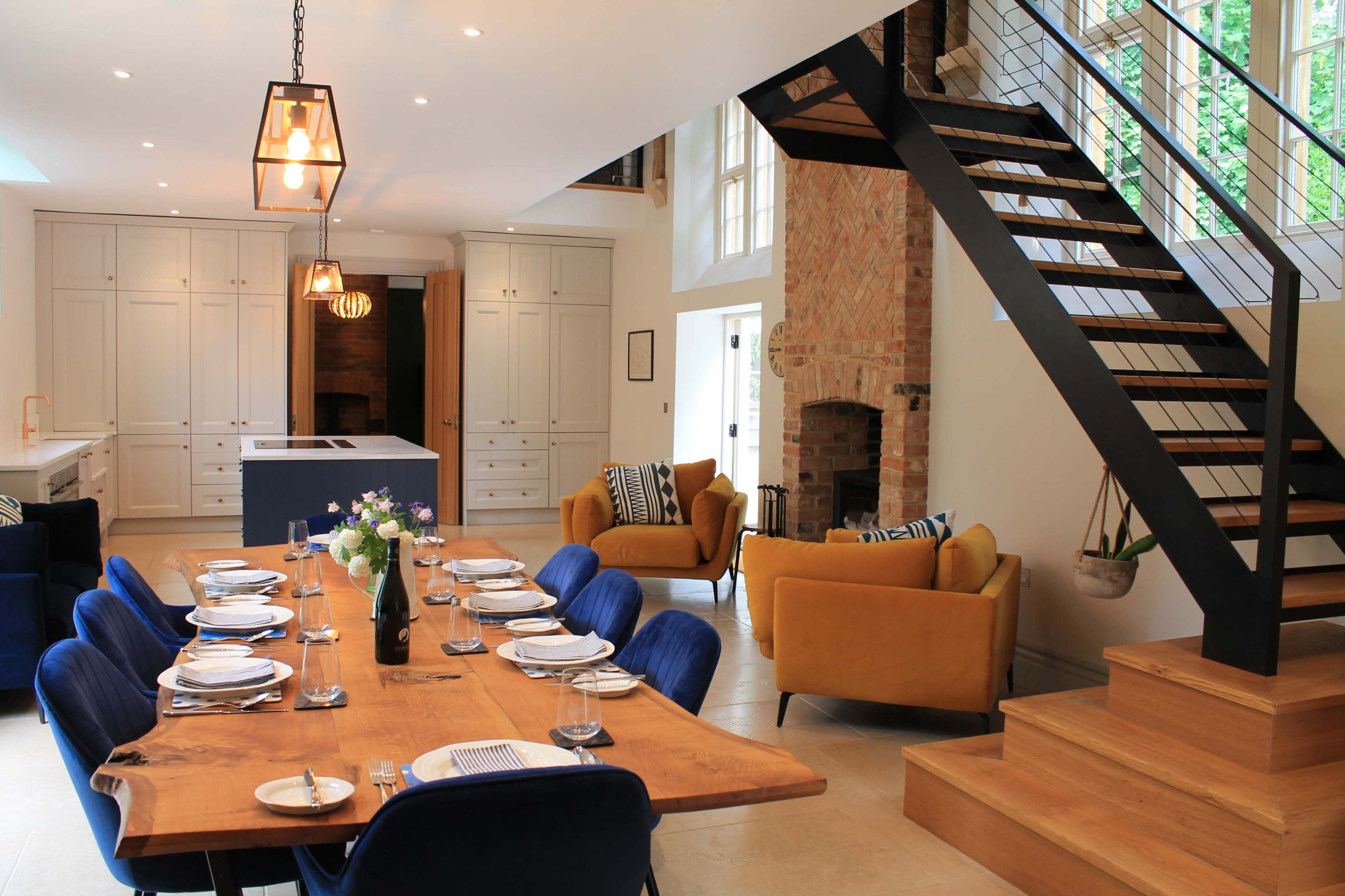 A modern dining room features a long wooden table set for a meal, with blue velvet chairs, yellow armchairs, and a staircase leading to an upper level.