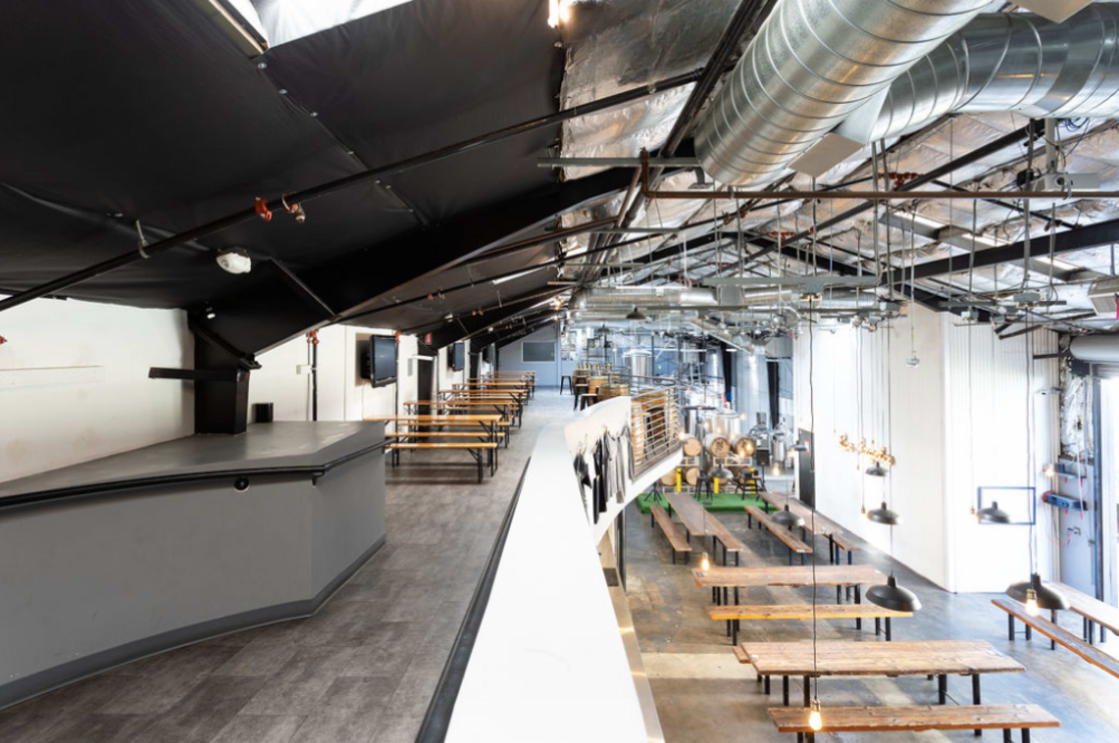 A spacious, modern brewery interior with a high ceiling, long communal tables, and visible brewing equipment in the background.