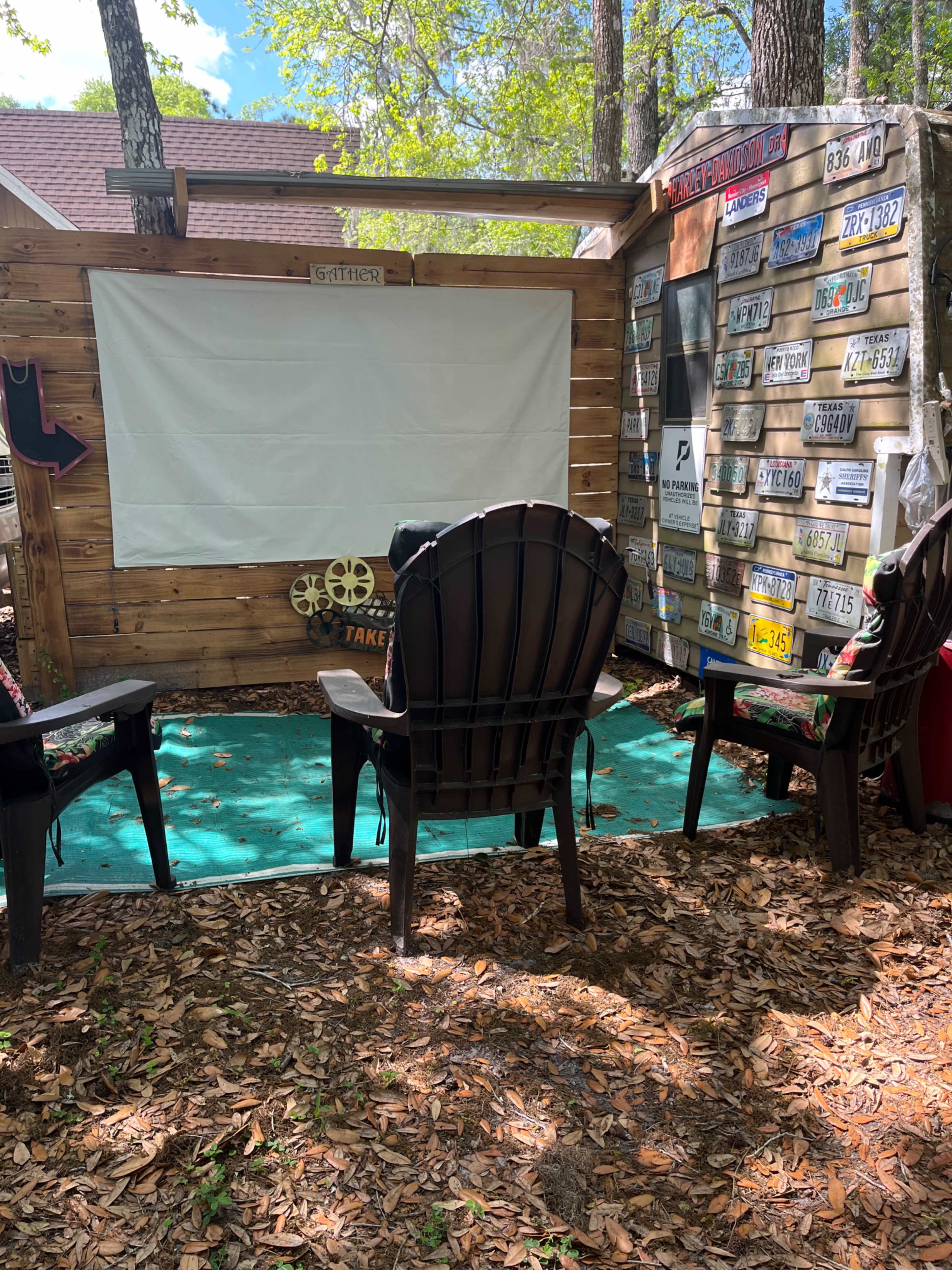 The scene features an outdoor sitting area with black adirondack chairs facing a white canvas screen, surrounded by a wooden backdrop and a wall adorned with various license plates.