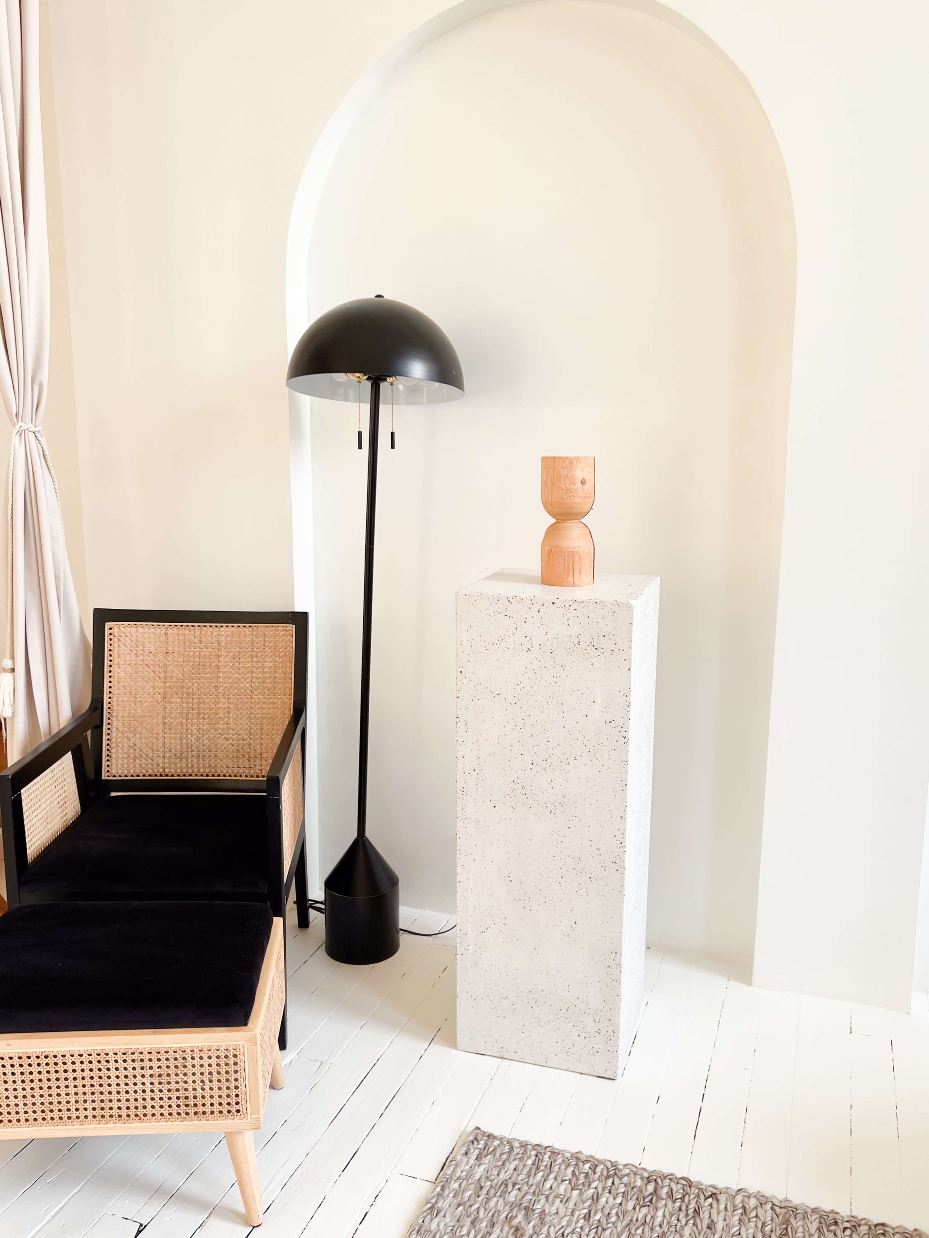 The image features a black and wood armchair next to a tall black floor lamp and a concrete pedestal displaying a wooden sculpture.