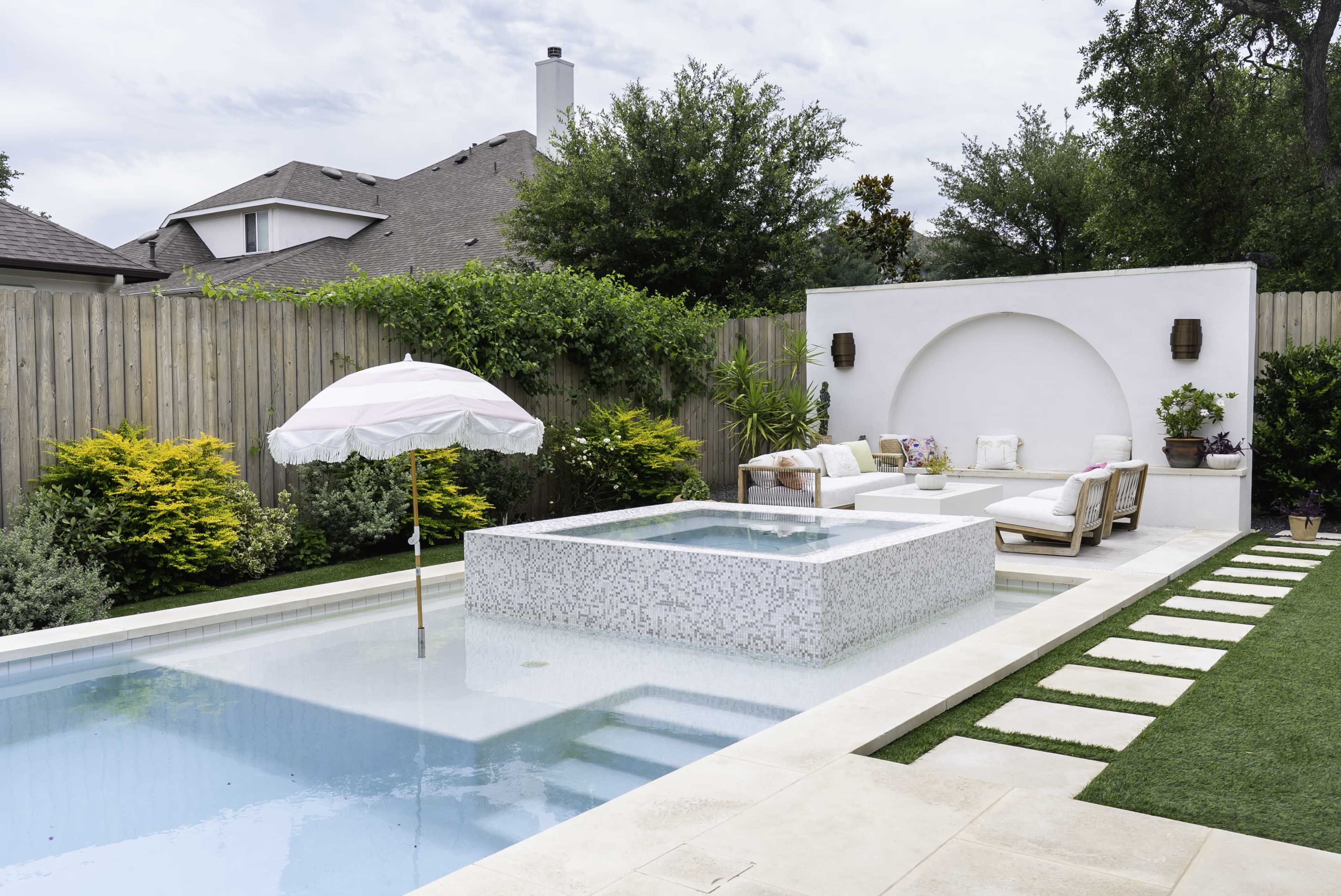 The image shows a landscaped backyard with a swimming pool that features a raised spa, an umbrella, and a seating area surrounded by greenery and wooden fencing.