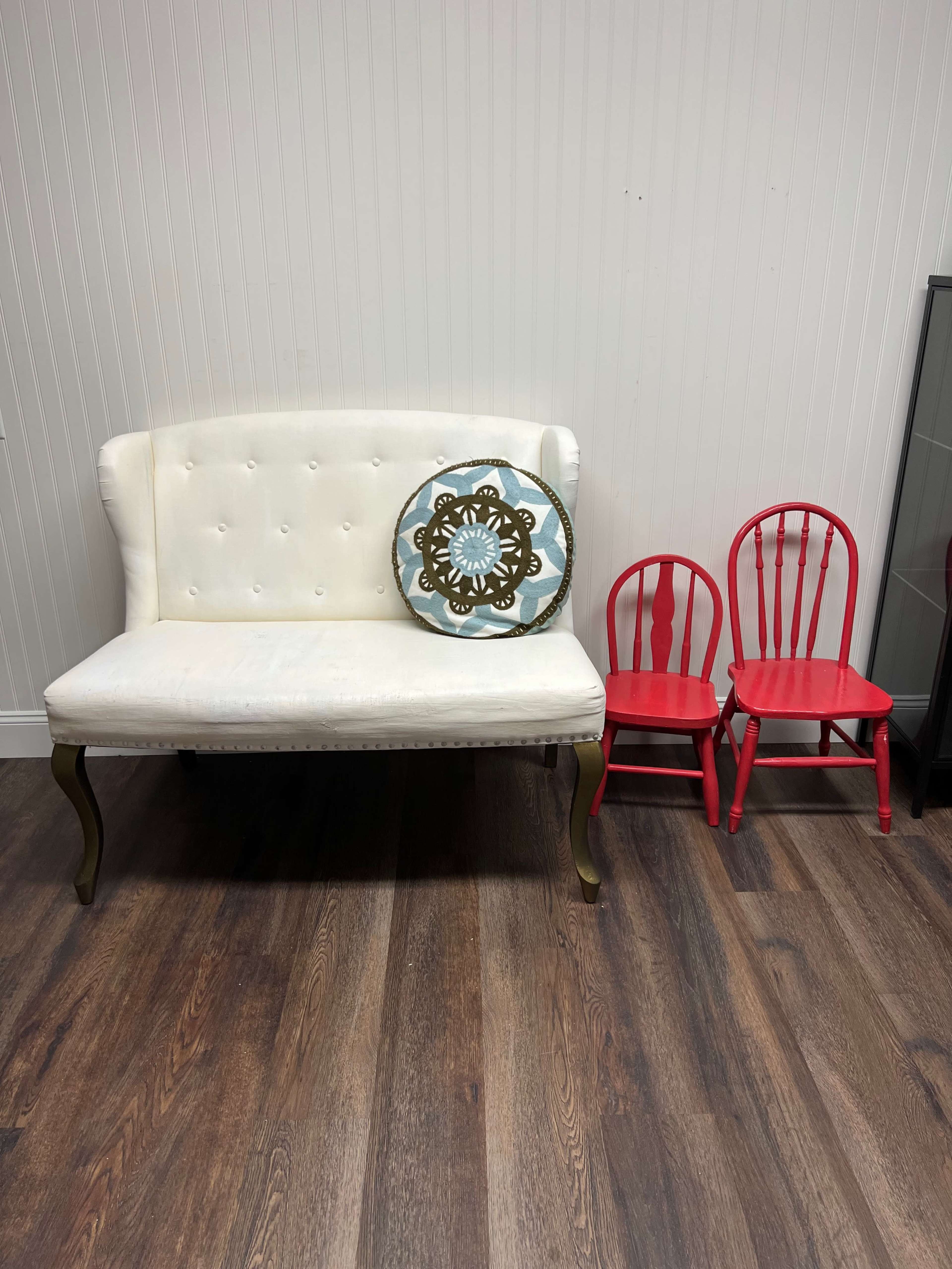 A white loveseat with a decorative blue and gold pillow is positioned next to two red children's chairs on a wood floor.