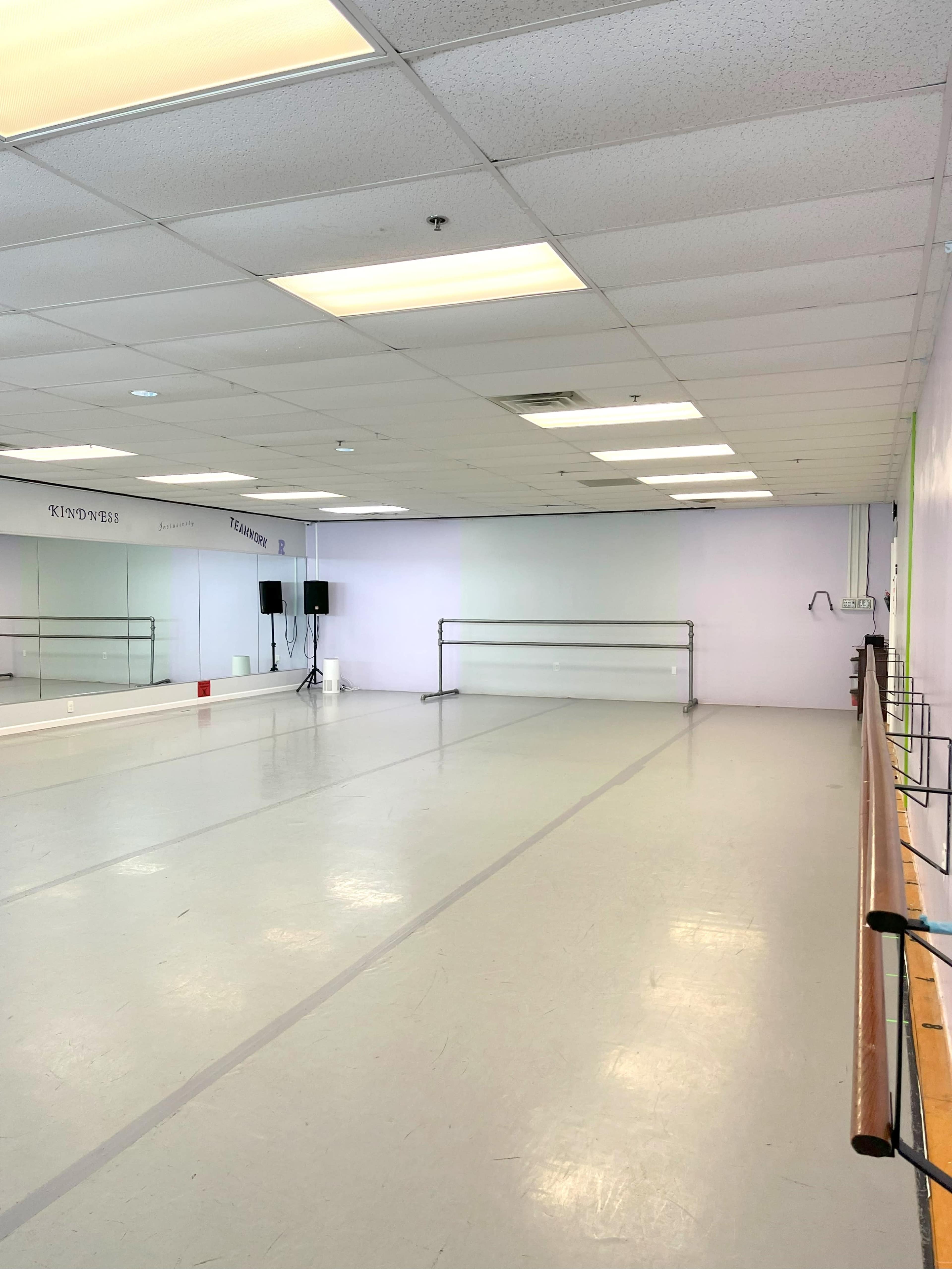 The image shows a spacious dance studio with mirrored walls, a wooden barre, and bright overhead lighting.