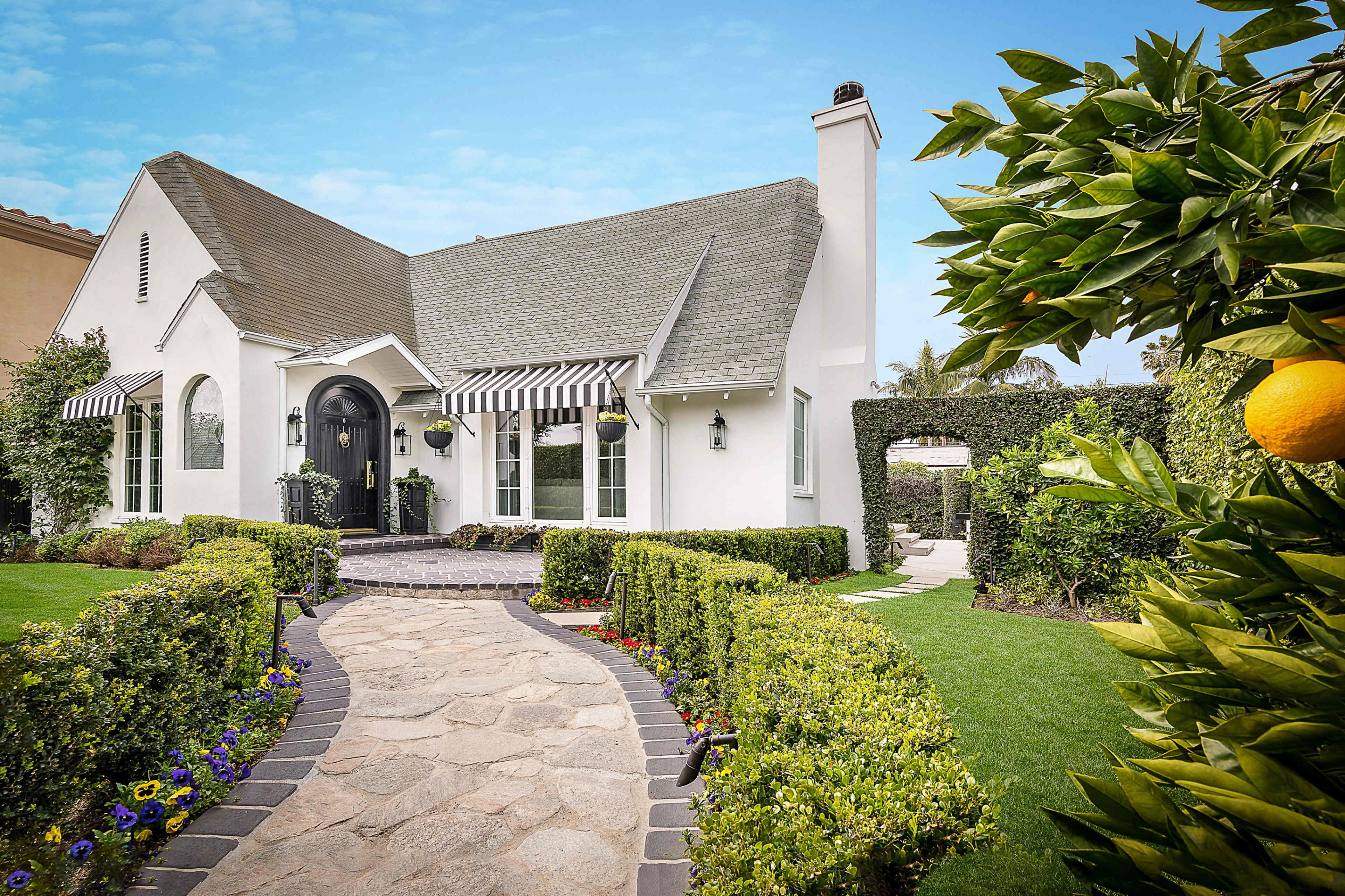 A white house with a grey roof and black door is surrounded by neatly trimmed hedges and a stone pathway leading to the entrance, with lemon trees visible on the right.