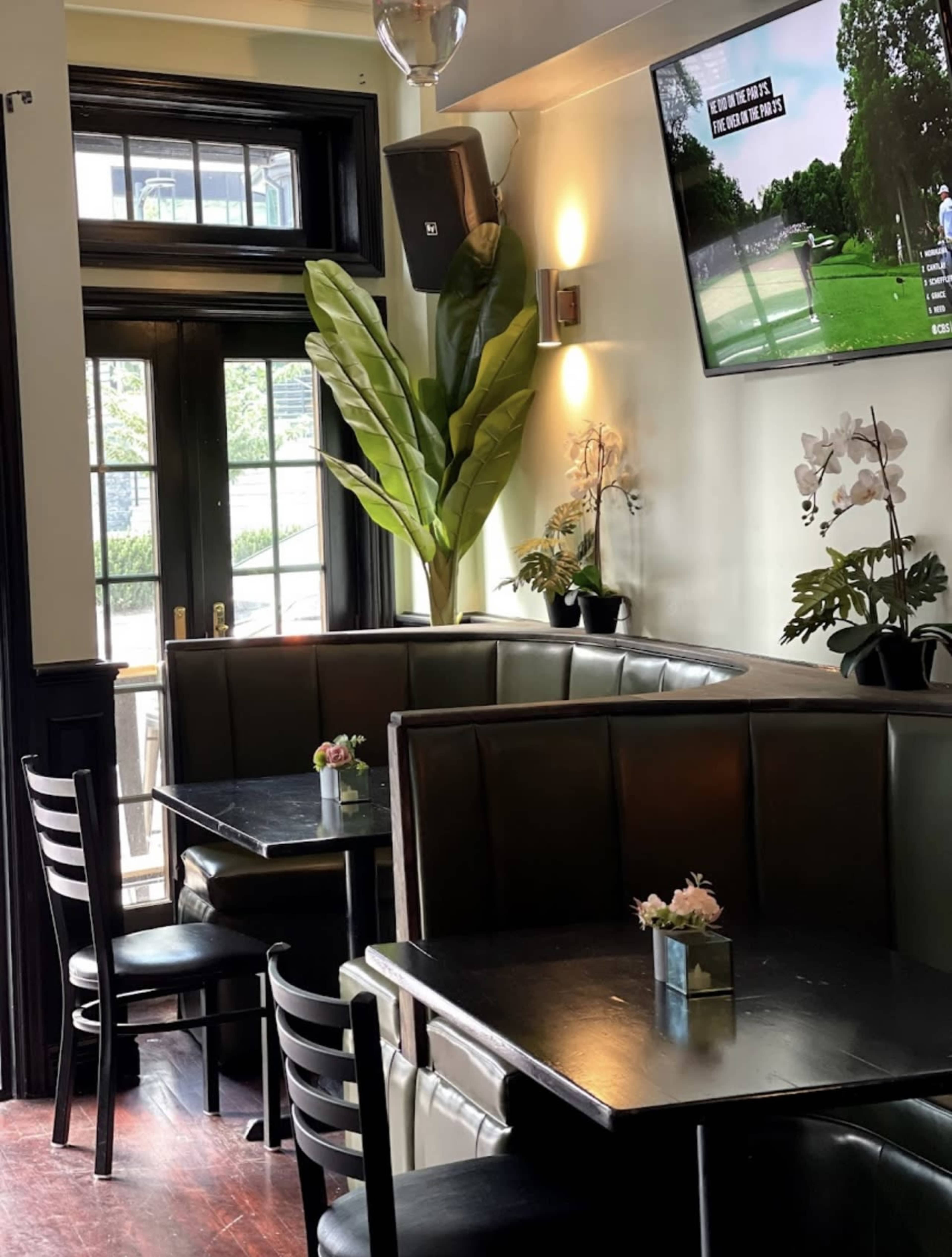The image shows a cozy restaurant interior with a dark booth, small tables, and a large window, featuring a TV mounted on the wall.