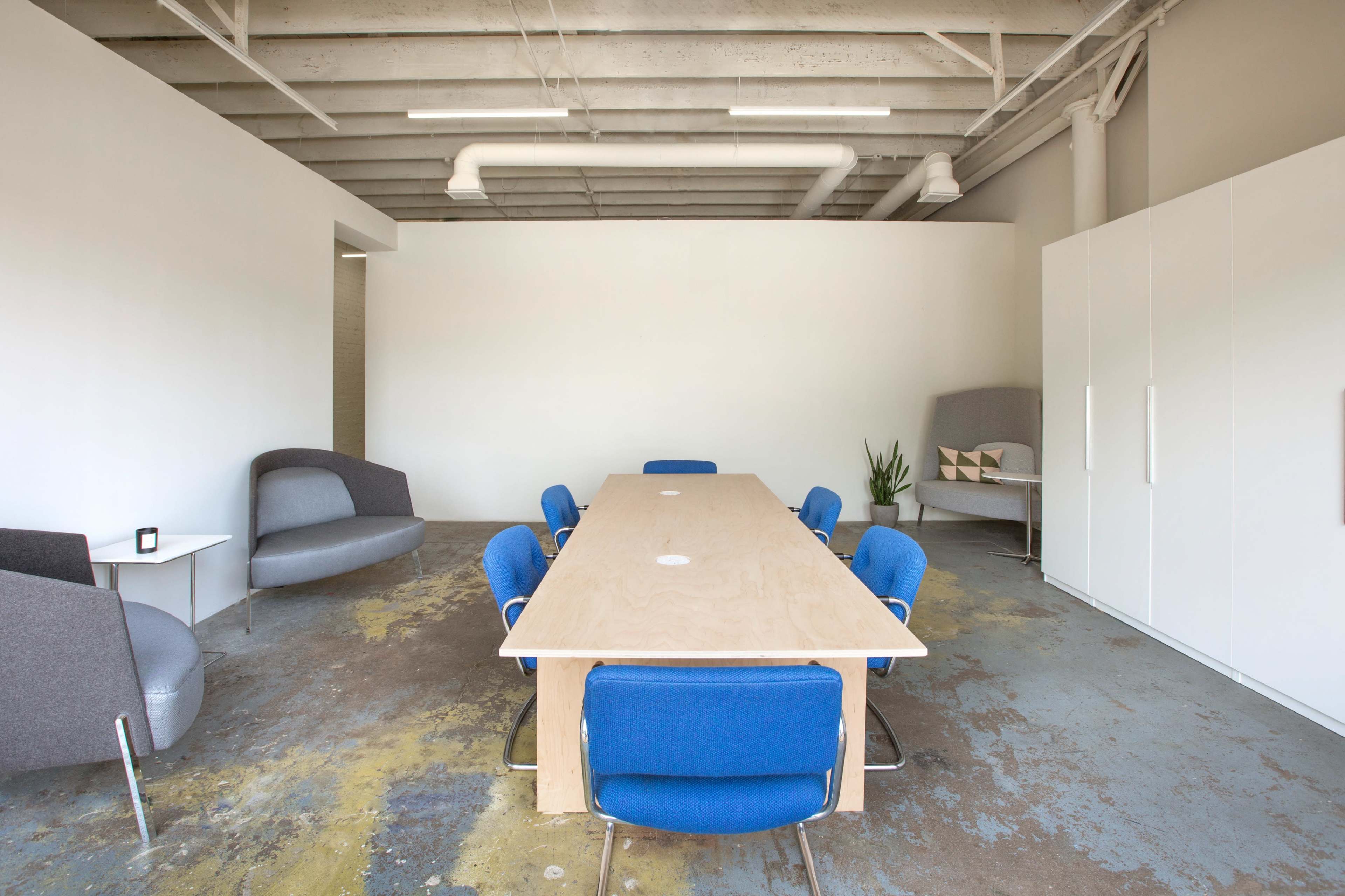 A long wooden table surrounded by blue chairs is the focal point of a spacious, minimalist conference room featuring gray seating and concrete flooring.