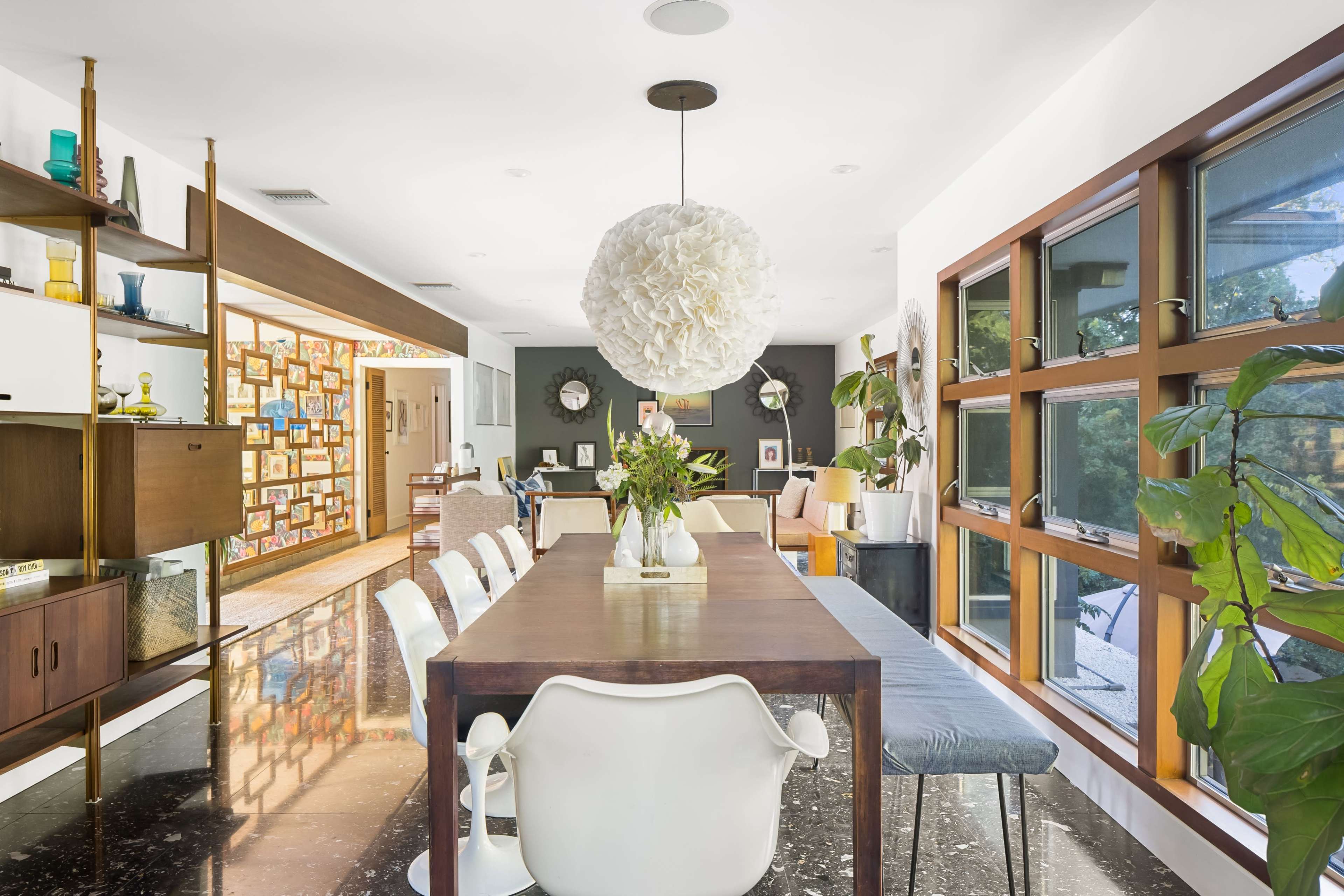 The image shows a modern dining area with a large wooden table surrounded by white chairs, a pendant light overhead, and large windows that provide natural light.