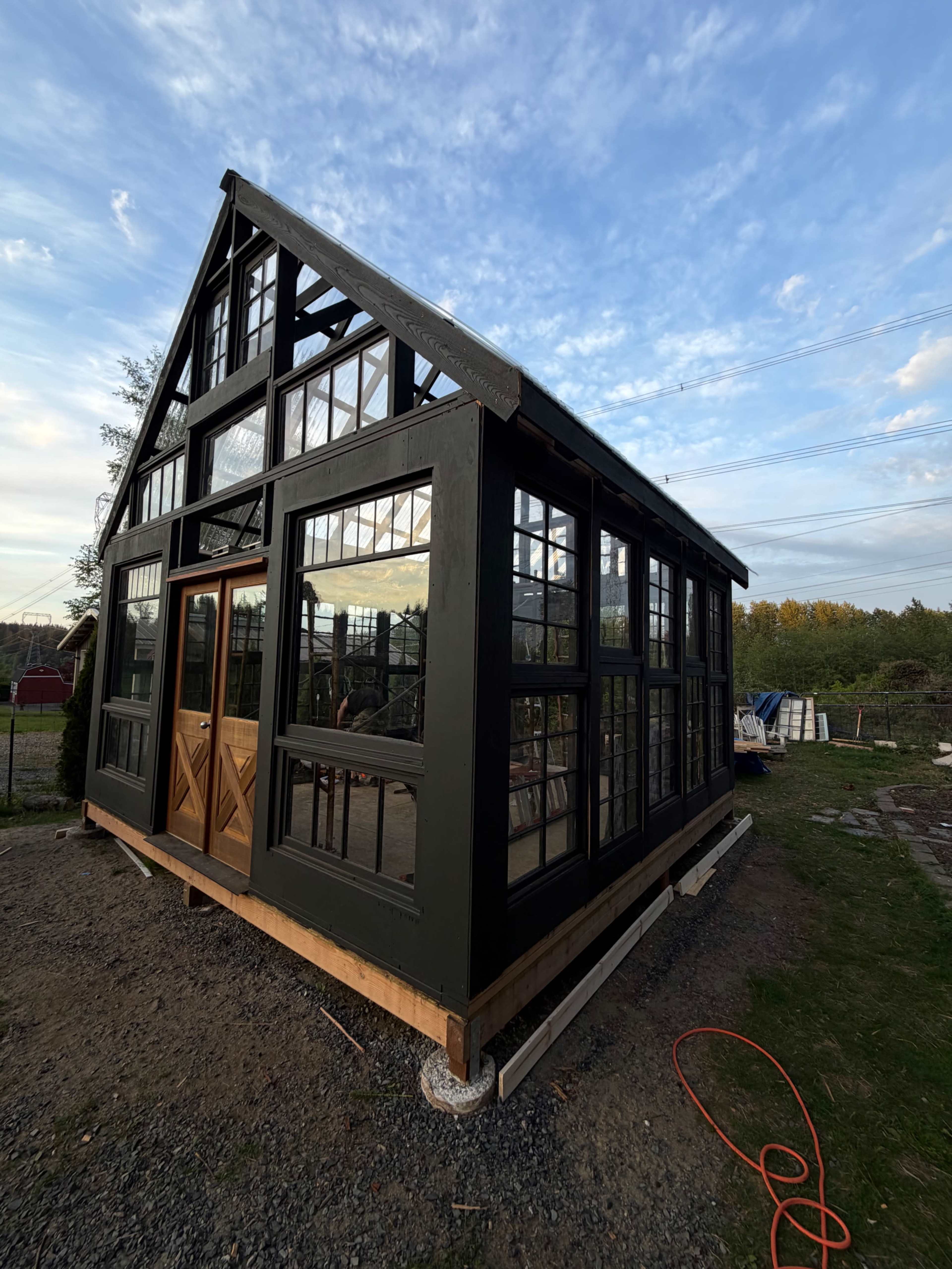 A black-framed greenhouse with large glass windows and a wooden door is situated on a gravel base, surrounded by greenery.