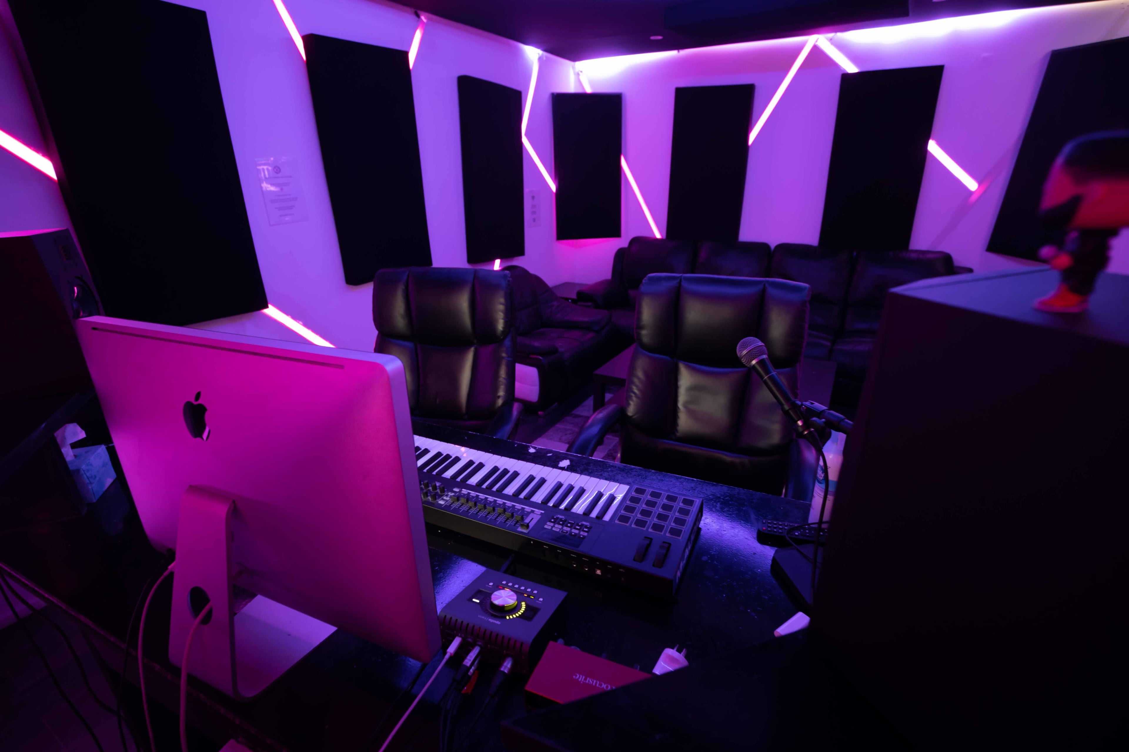 A modern recording studio with black leather chairs, a computer with a music keyboard, and vibrant neon lights along the walls.